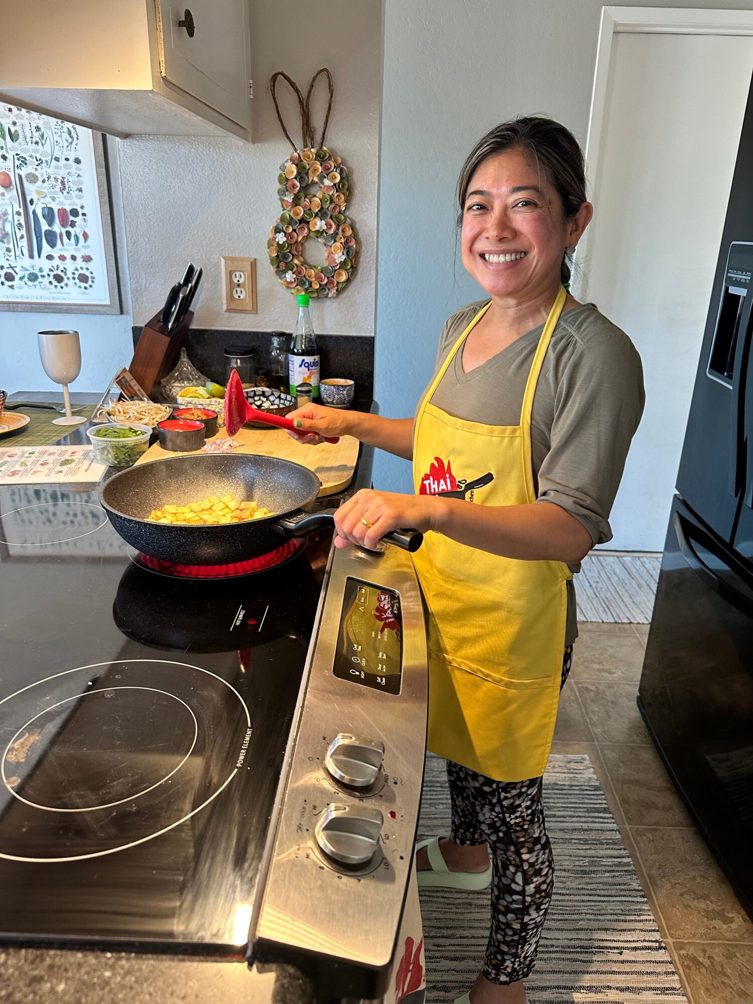 Woman cooking in kitchen, wearing yellow apron, smiling, with stove and kitchen decor visible.