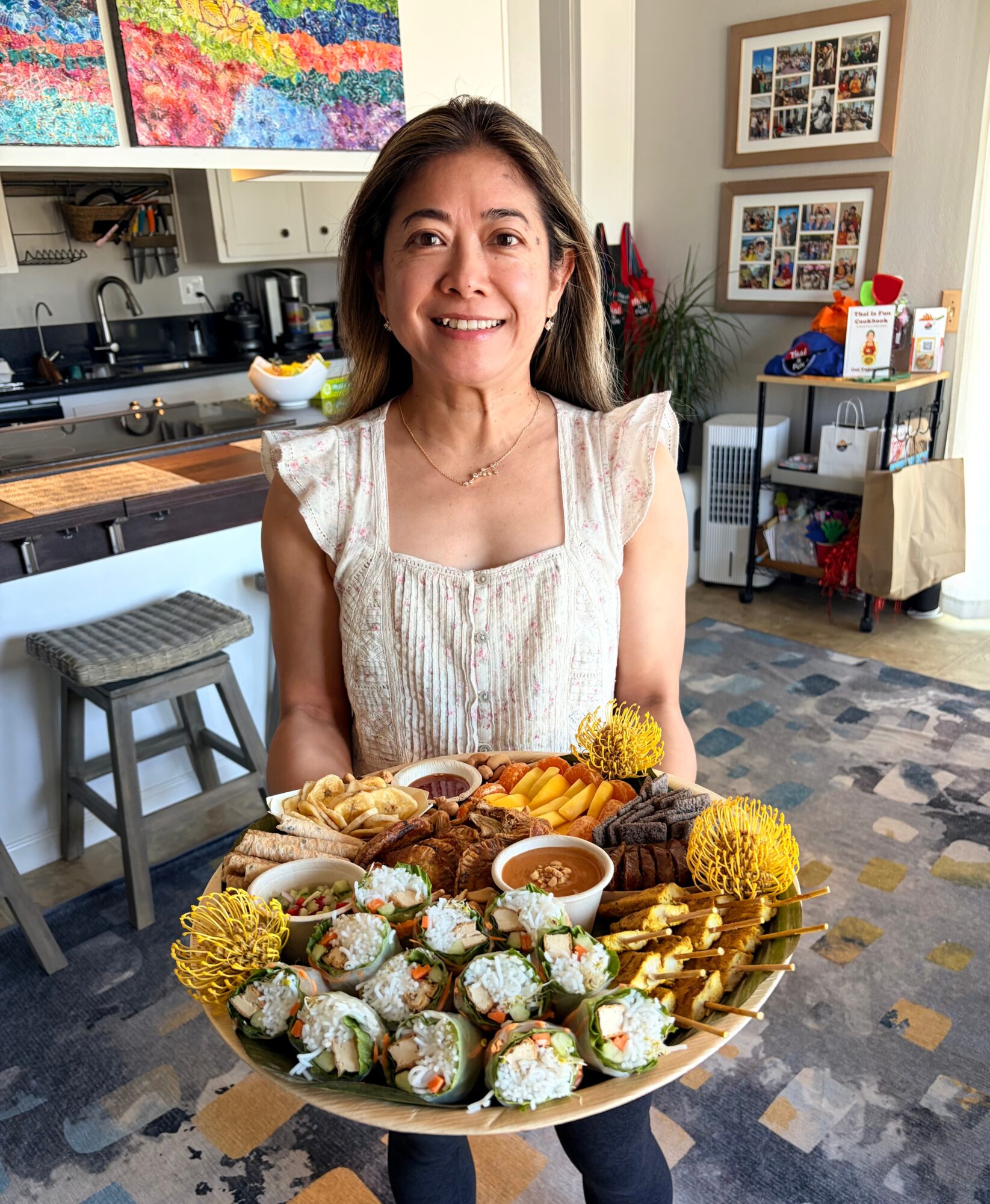 Woman holding a large tray of assorted sushi and fried snacks, smiling in a cozy kitchen or cafe setting.