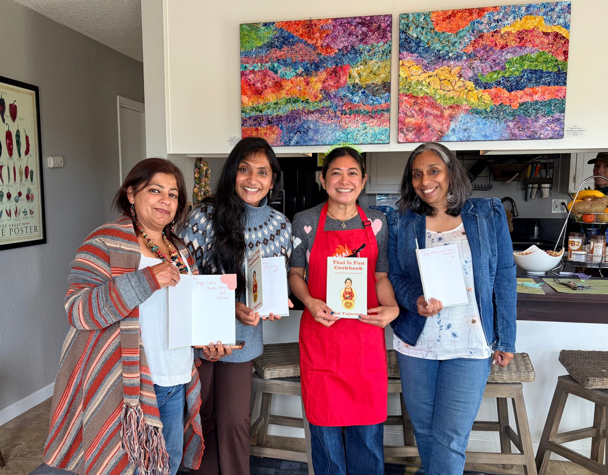 Four women stand together indoors, holding gift bags and a cookbook, with colorful abstract paintings on the wall behind them.