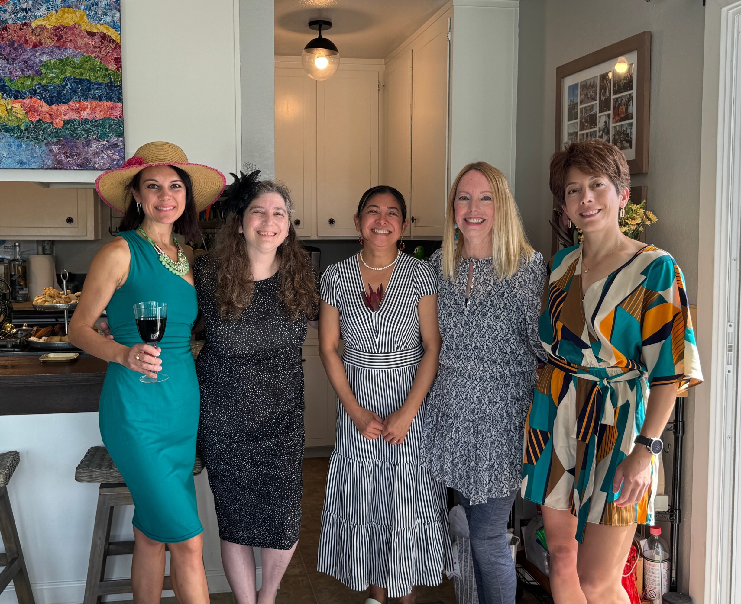 Five women standing together indoors, smiling, with one holding a glass of wine, in a home setting.
