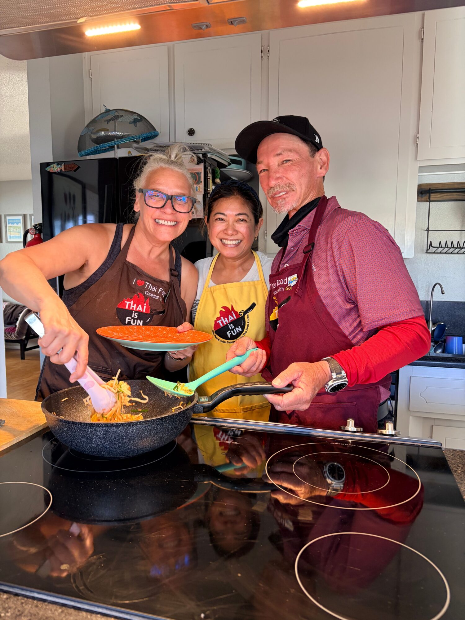 Three people cooking together in a kitchen, one stirring a pot on the stove, others smiling and holding utensils.