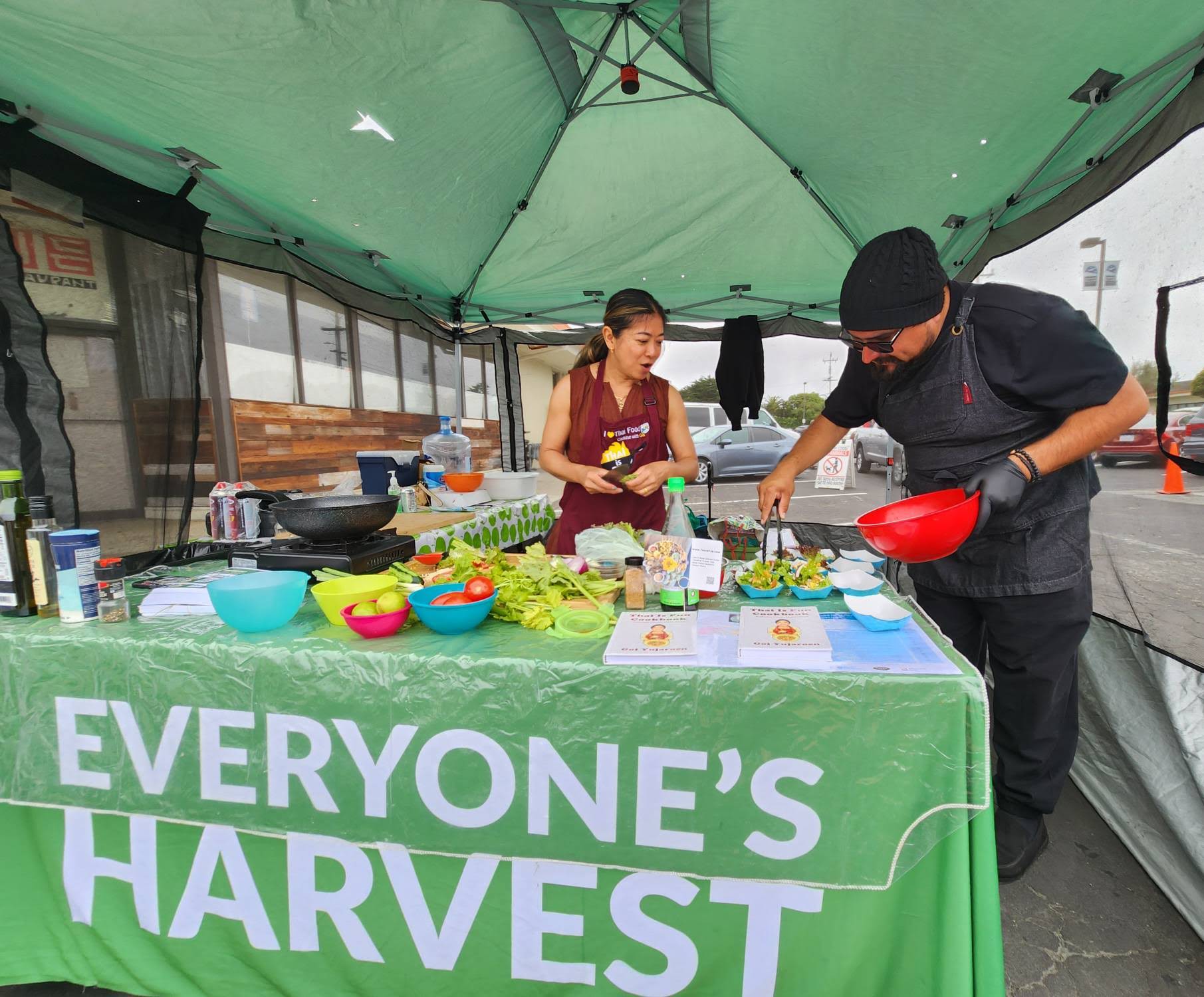 Two people preparing food at a harvest event under a green tent, with a table labeled 'Everyone's Harvest' and various vegetables and bowls.