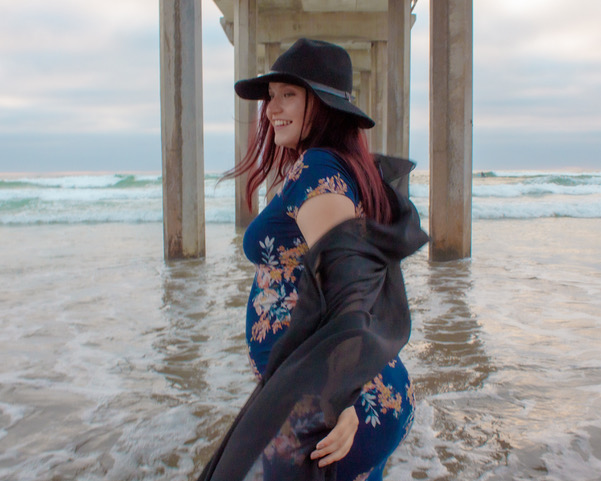 Woman in a floral dress and hat smiling on the beach near a pier, waves in the background.
