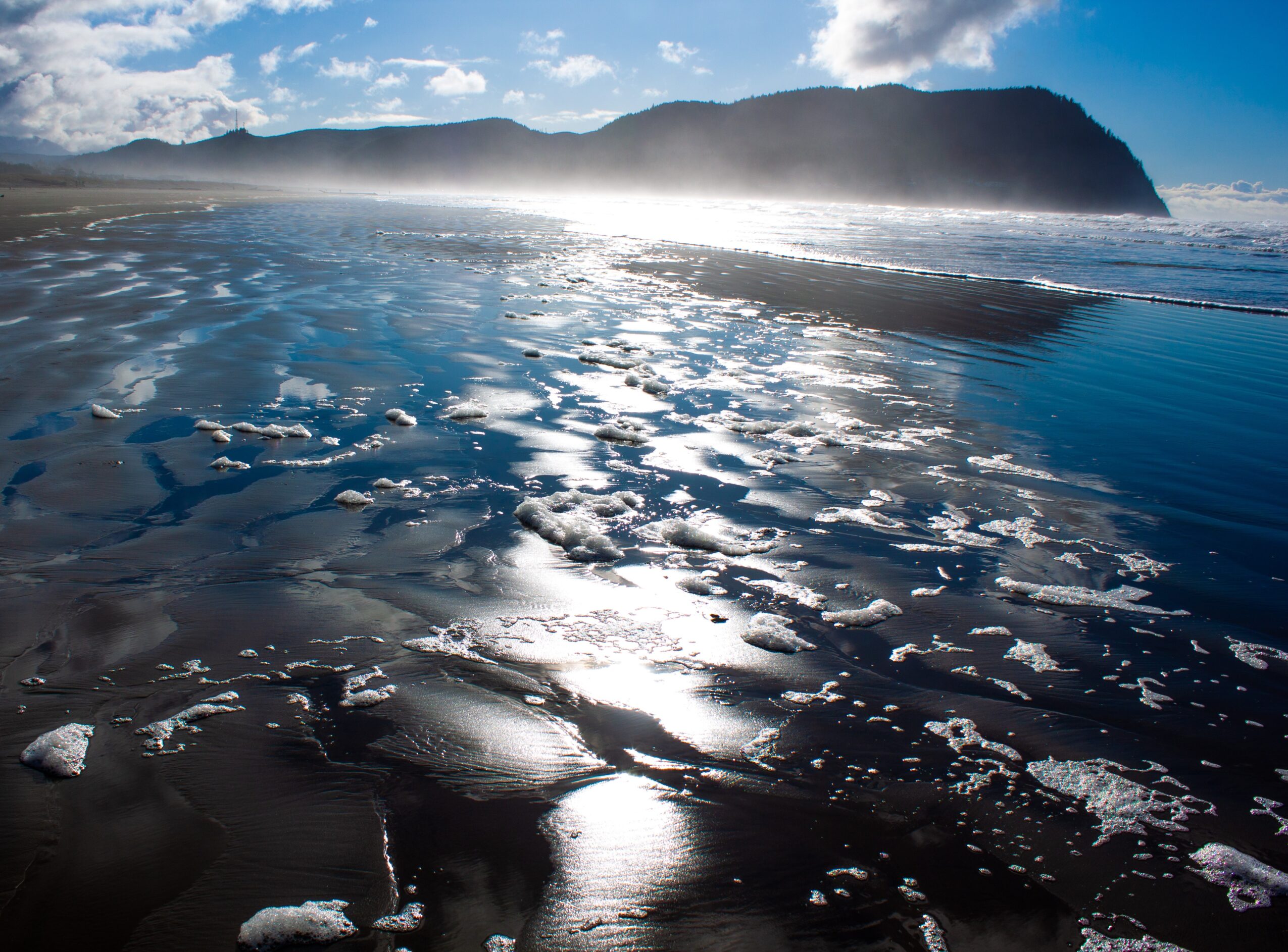 Ocean waves with foam and reflections, mountain in background under partly cloudy sky.