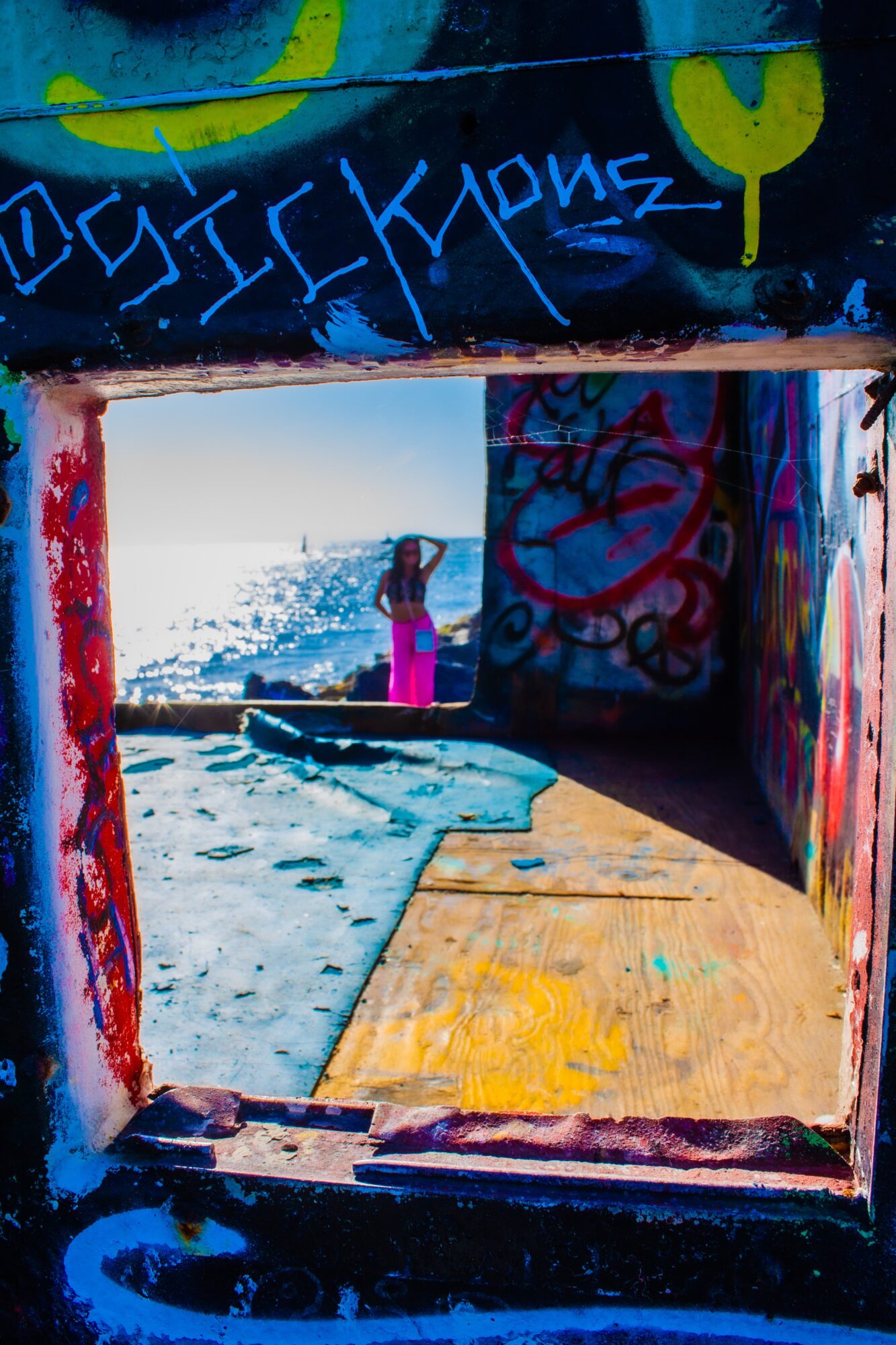 Person standing on beach near water, viewed through a graffiti-covered opening, with a bright sky and ocean in background.