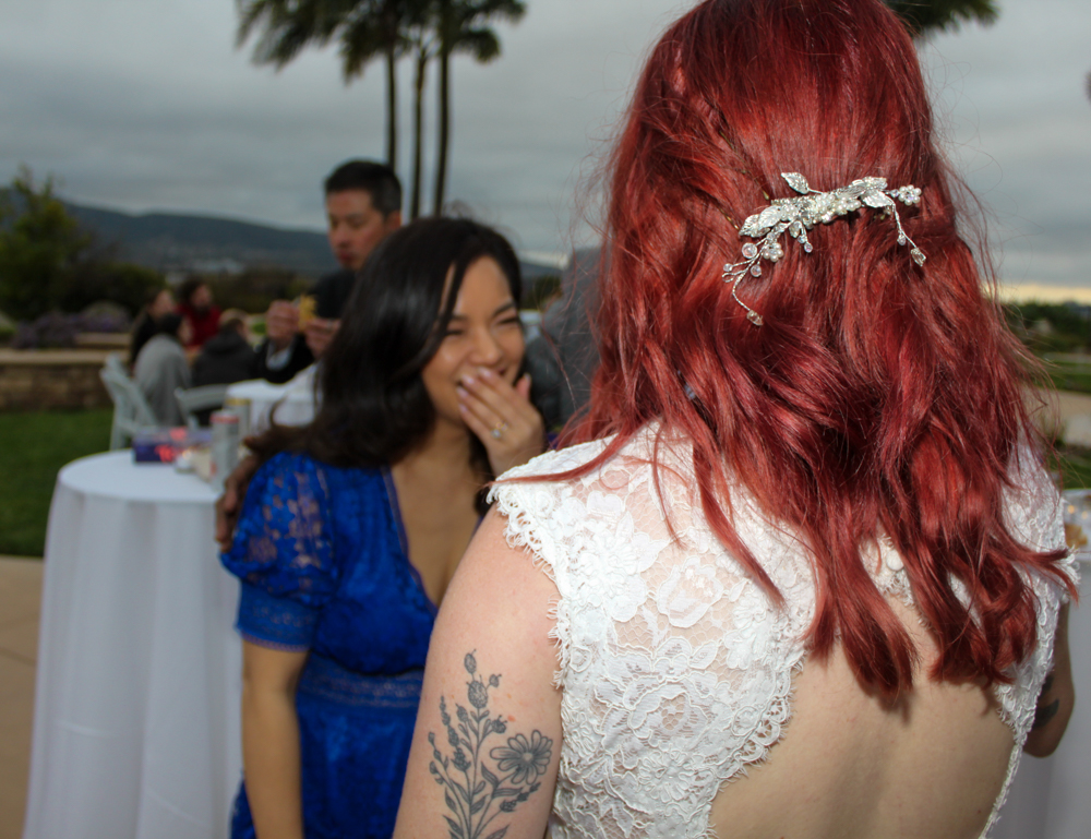 Two women at an outdoor event, one with red hair and a floral hair accessory, the other with black hair, smiling and talking.