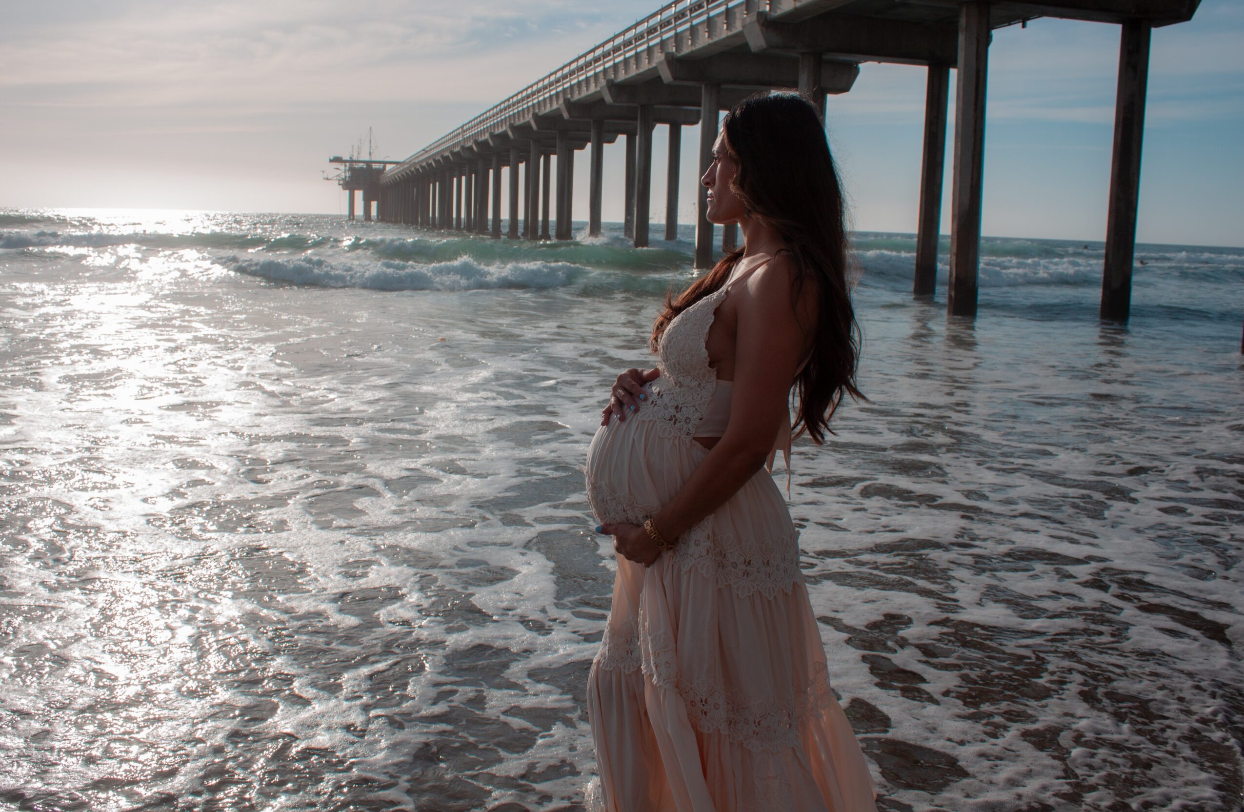 Pregnant woman in a flowing dress stands on the beach near a pier, with ocean waves and sunlight in the background.
