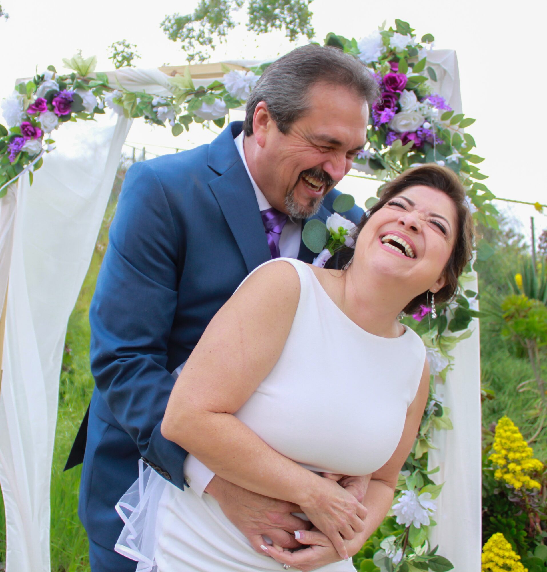 A smiling couple, a man in a blue suit and a woman in a white dress, embrace outdoors near a floral arch.