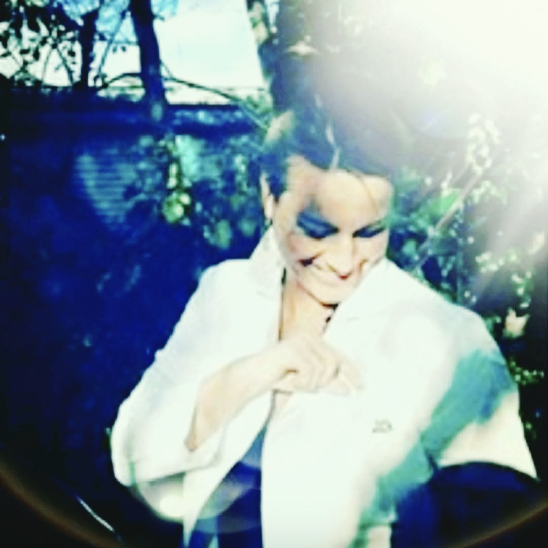 Person with short hair and makeup, wearing a white jacket, smiling outdoors with sunlight and trees.