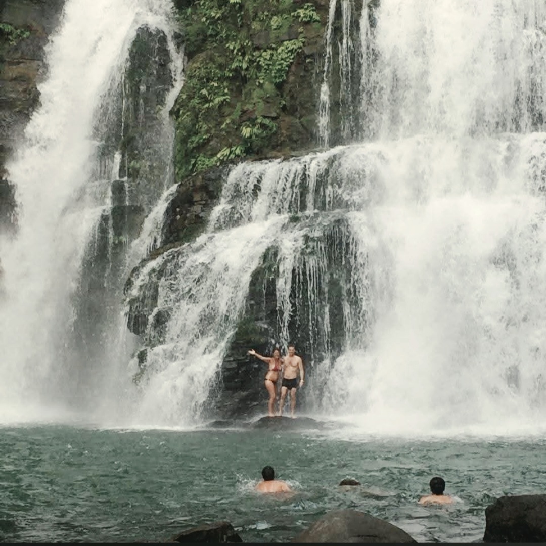 Two people standing under a waterfall, with three others swimming nearby in a pool, surrounded by rocks and greenery.