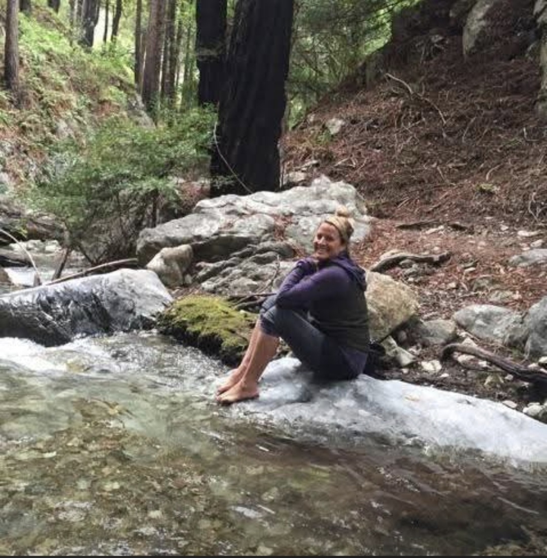 Person sitting on a rock by a stream in a forest, smiling, with legs in the water, surrounded by trees and rocks.