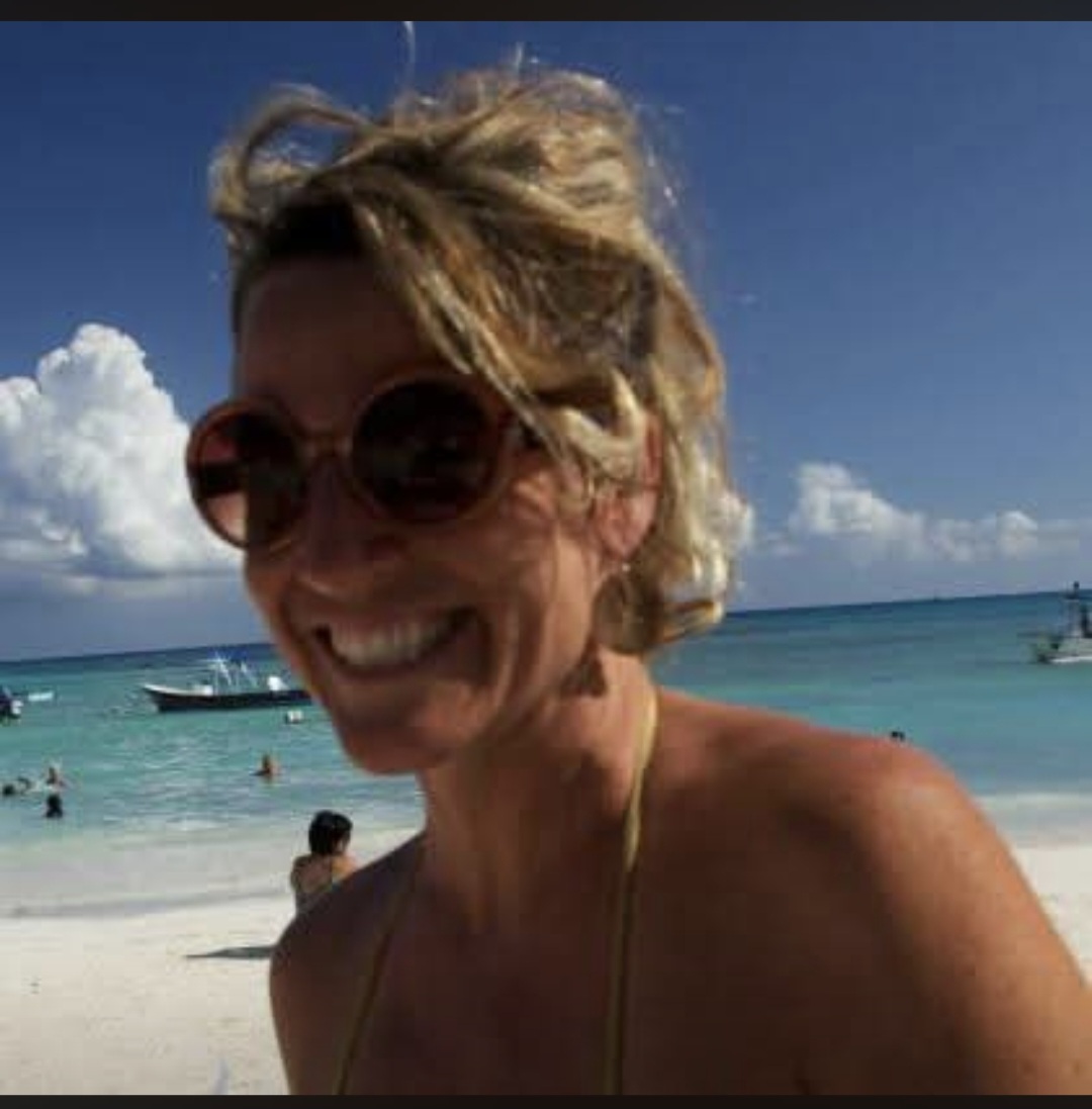 Smiling woman with sunglasses at the beach, ocean and boats in the background, sunny day.