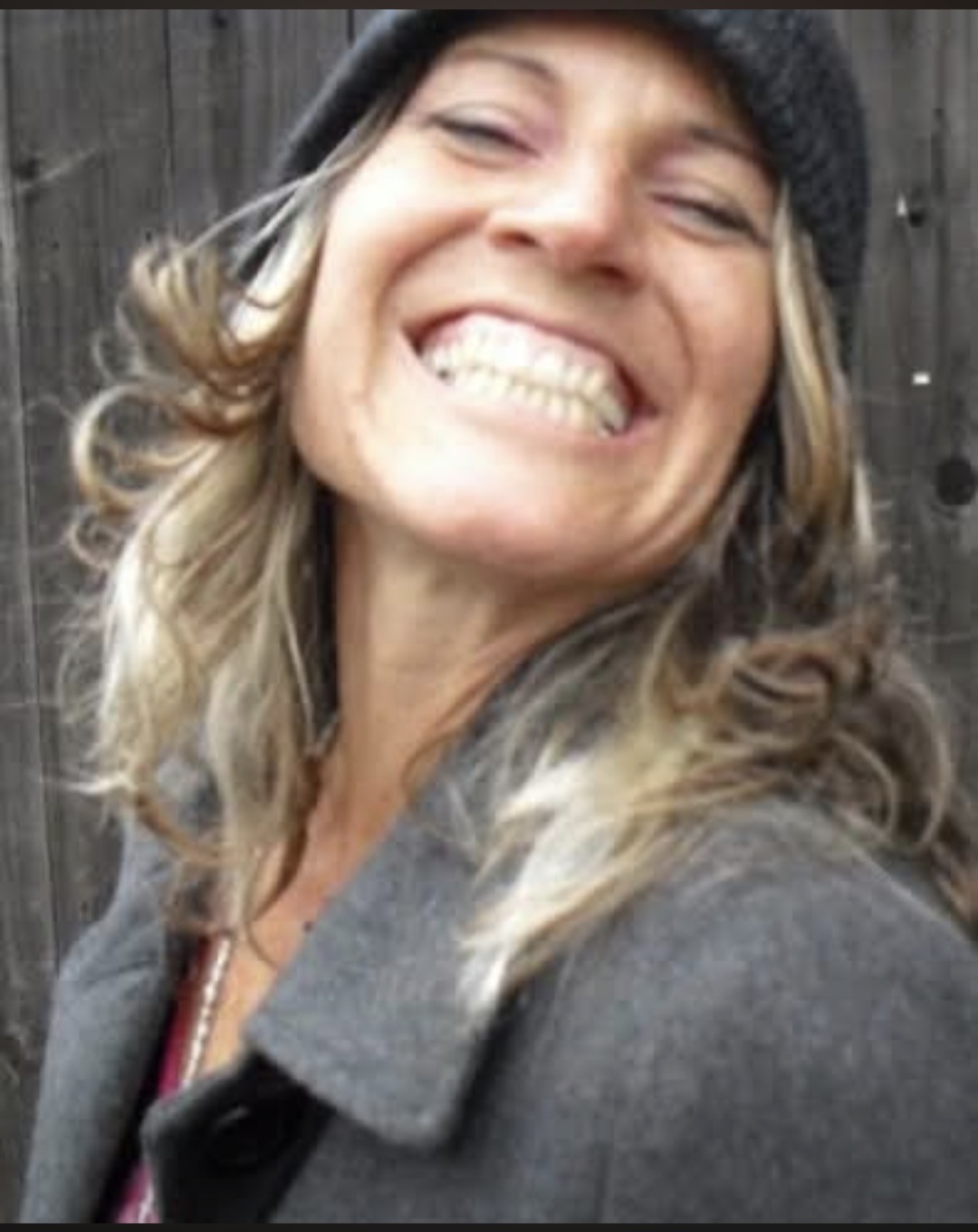 Woman smiling broadly, wearing a black beanie and gray coat, outdoors with a wooden fence background.