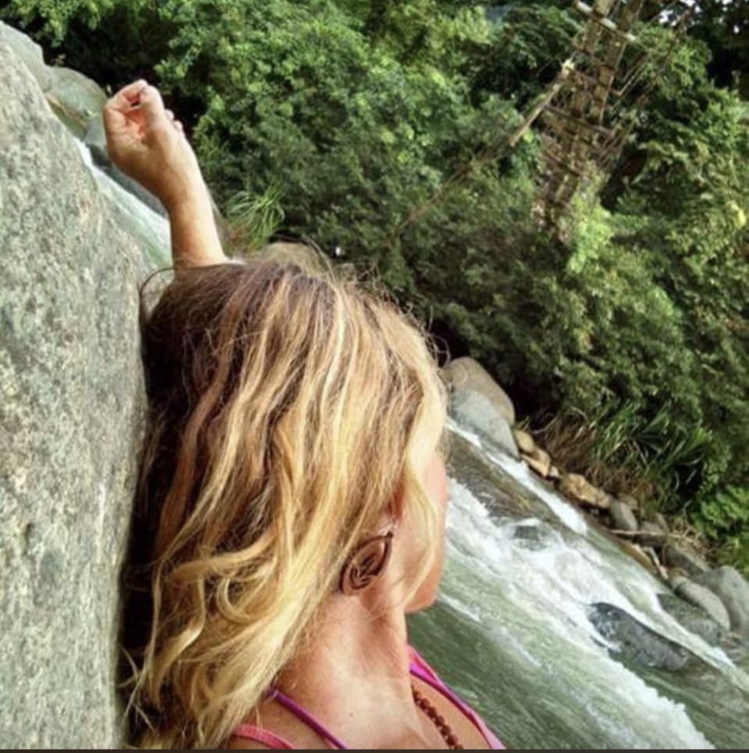 Woman with long blonde hair leaning against a rock near a river, surrounded by trees and greenery.