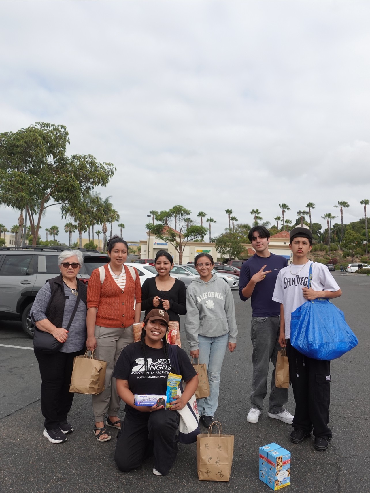 Group of seven people standing outdoors with trees and parking lot in background, some holding bags and boxes, overcast sky.