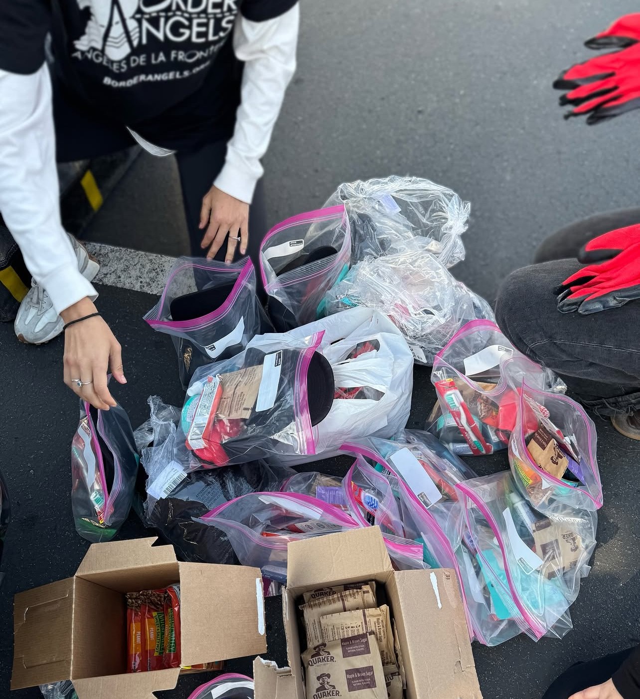 People are organizing and packing items into clear plastic bags on a table, with some boxes nearby, on a gray surface.
