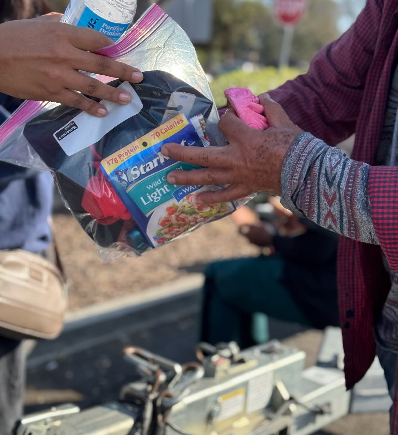 Two people exchange a plastic bag containing food and supplies outdoors, with a blurred background and a person sitting nearby.