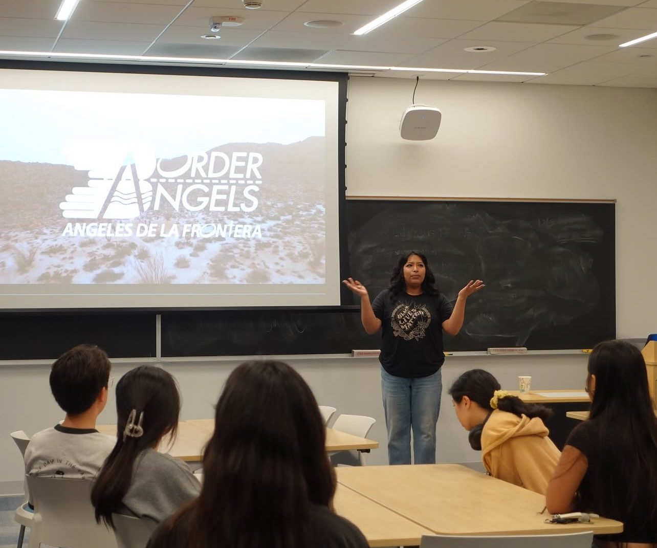 A woman stands in front of a classroom, gesturing with her hands, with students seated at desks and a presentation slide behind her.