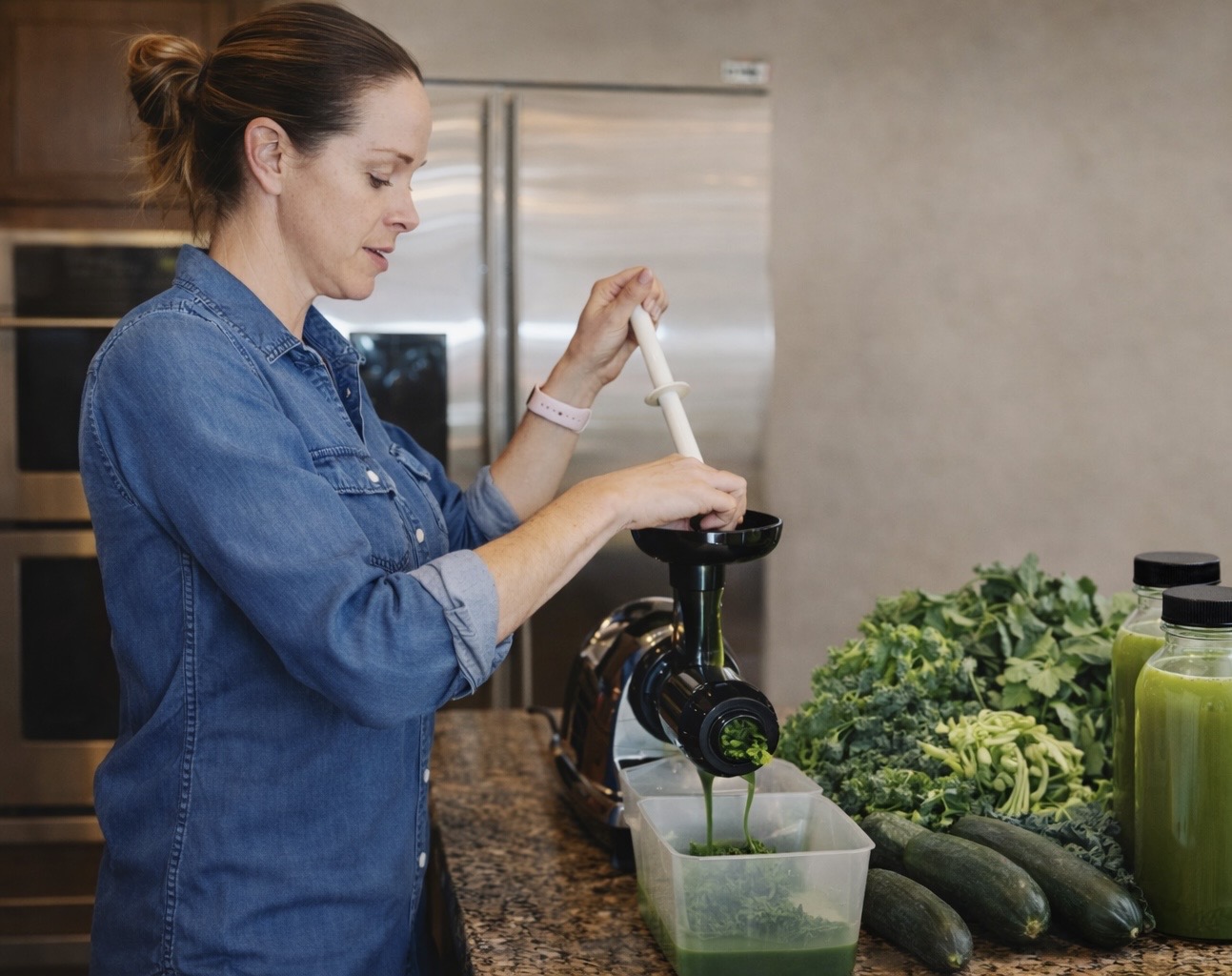 Woman in denim shirt using a juicer with green vegetables and bottles nearby, in a kitchen setting.