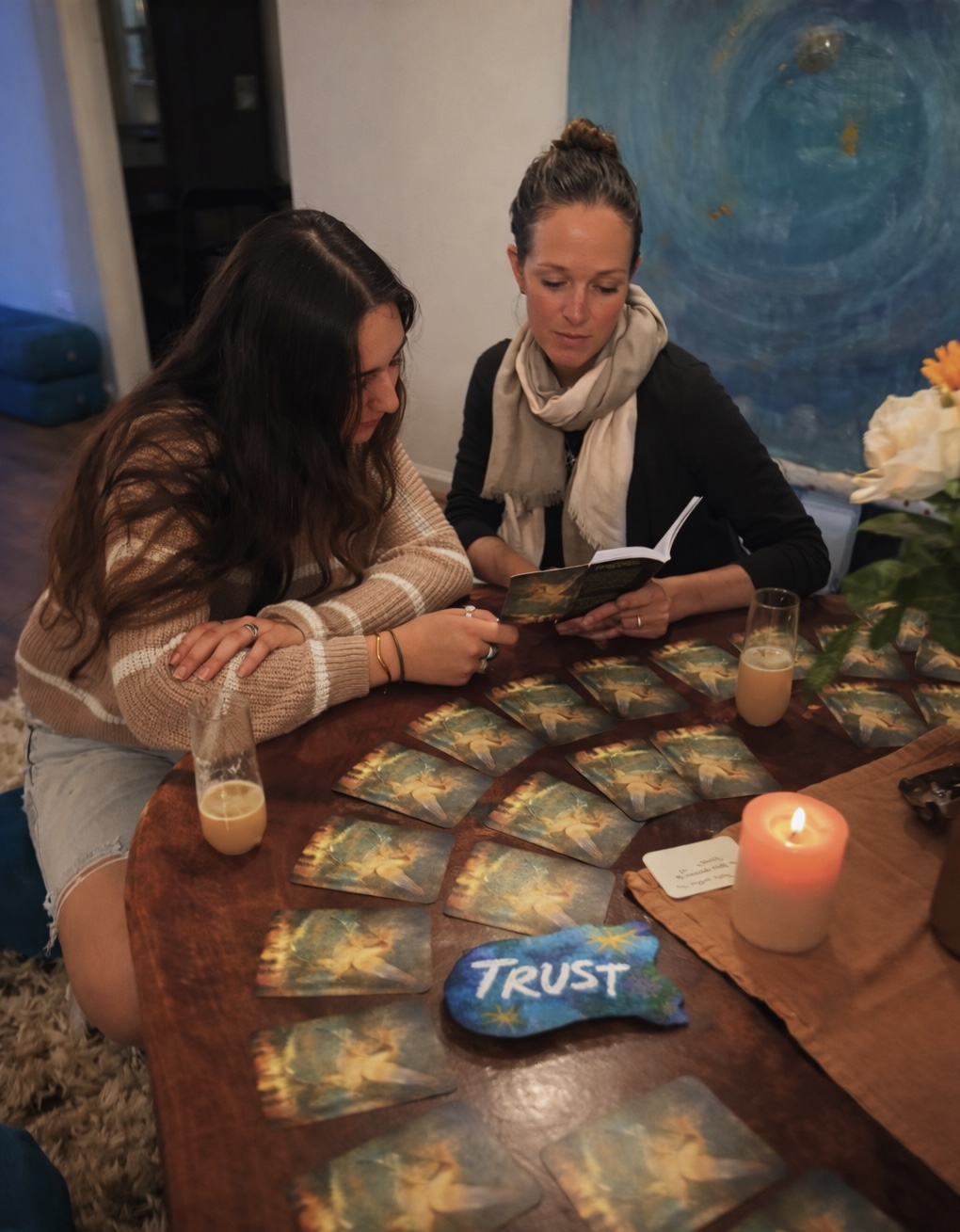 Two women sit at a table with tarot cards, candles, and flowers, reading a book together indoors. A blue painting is in the background.