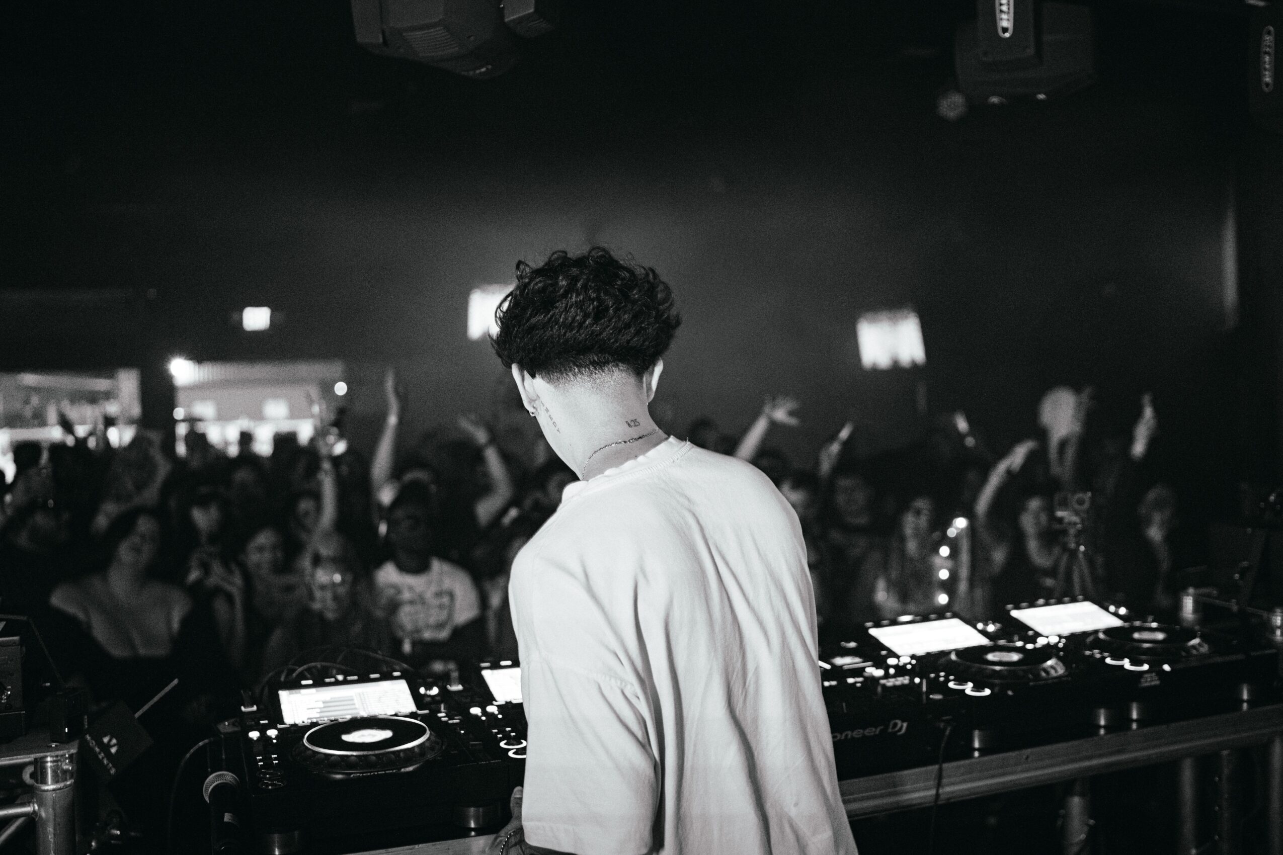 Person with curly hair facing a DJ booth in a crowded nightclub, with people dancing and raising hands.