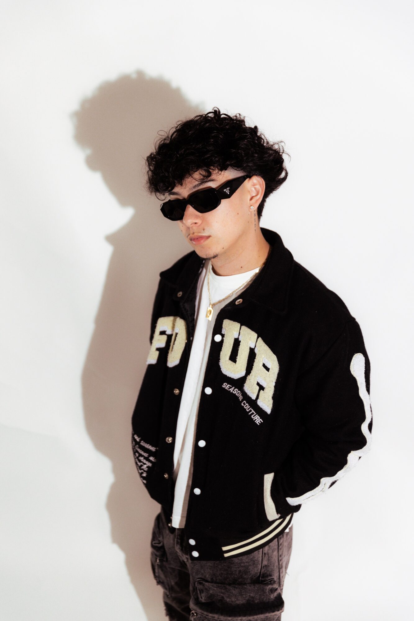 Young man with curly hair wearing sunglasses, a black varsity jacket, and a white shirt, standing against a white background.