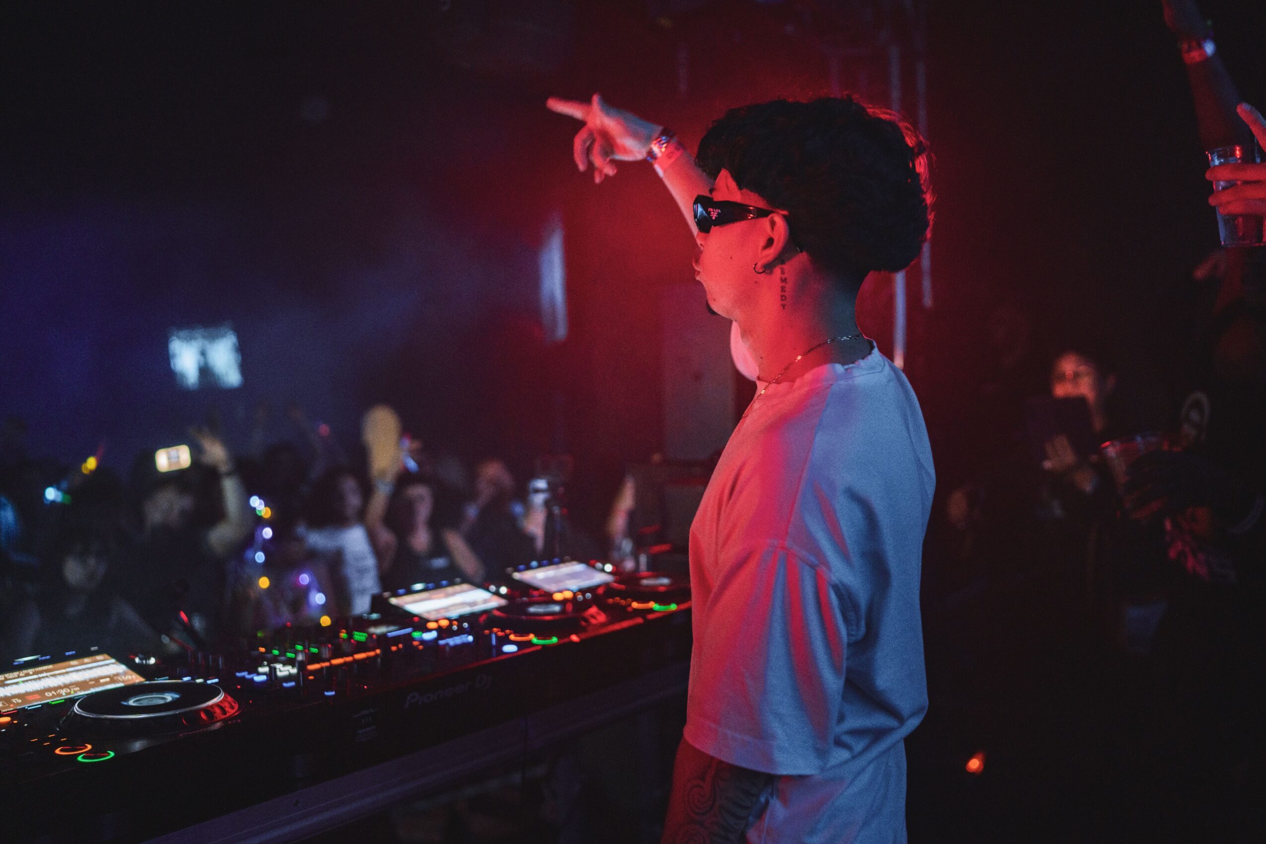 Person with short hair and glasses points at a DJ booth in a dark, colorful club setting with a crowd.