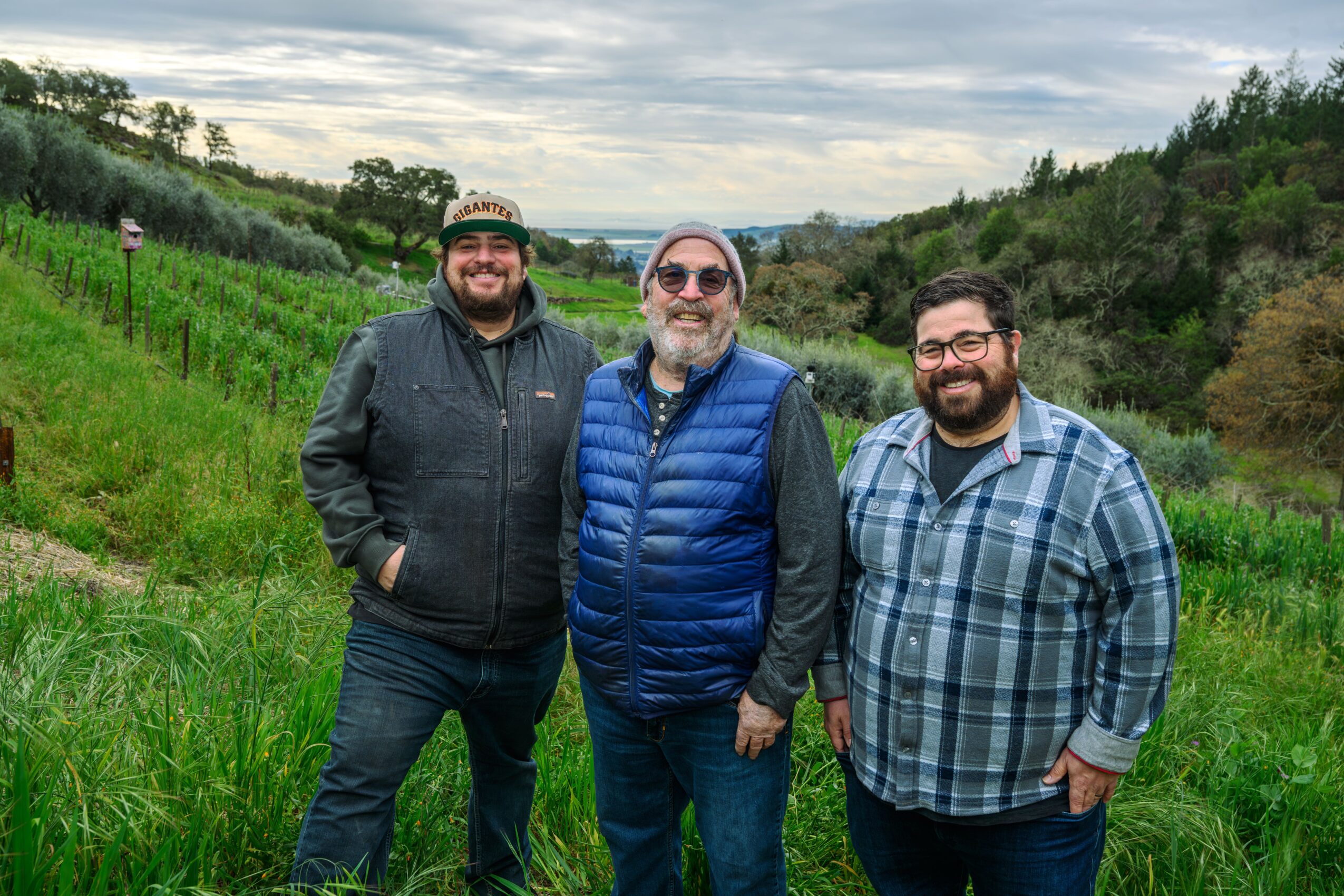 Three men standing outdoors in a green landscape with trees and hills, smiling at the camera.