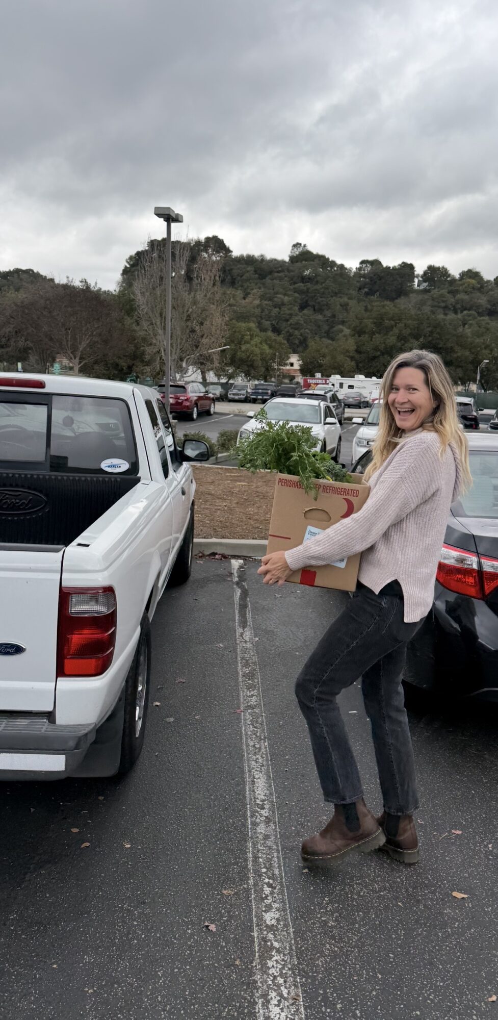 Woman smiling and holding a cardboard box in a parking lot with cars and a hill in the background.