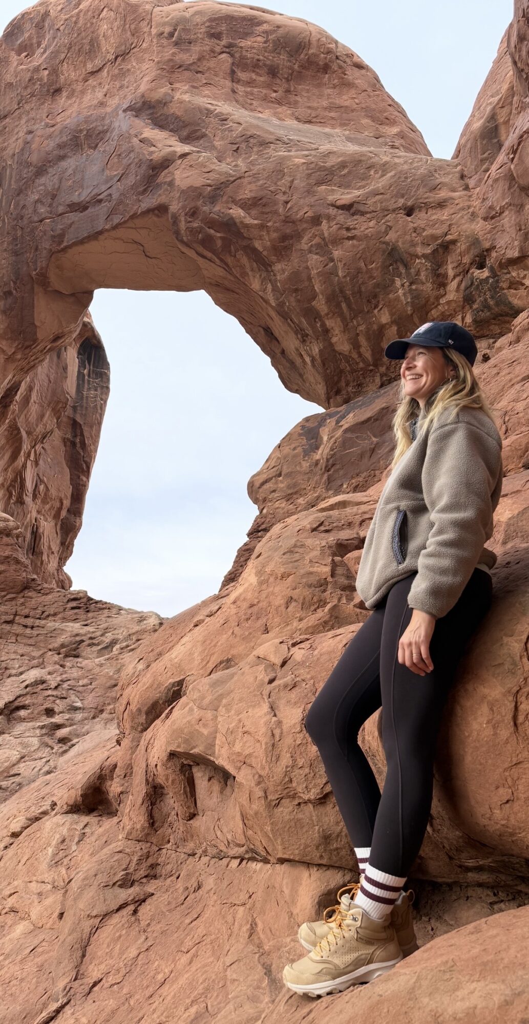Person standing near a natural rock formation with a heart-shaped opening, wearing a cap, jacket, and sneakers.