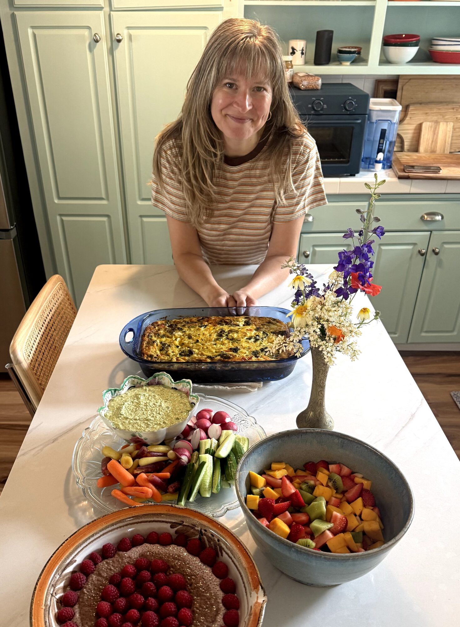 Woman with long hair smiling at a table with a casserole, salad, fruit, and a flower vase in a kitchen.