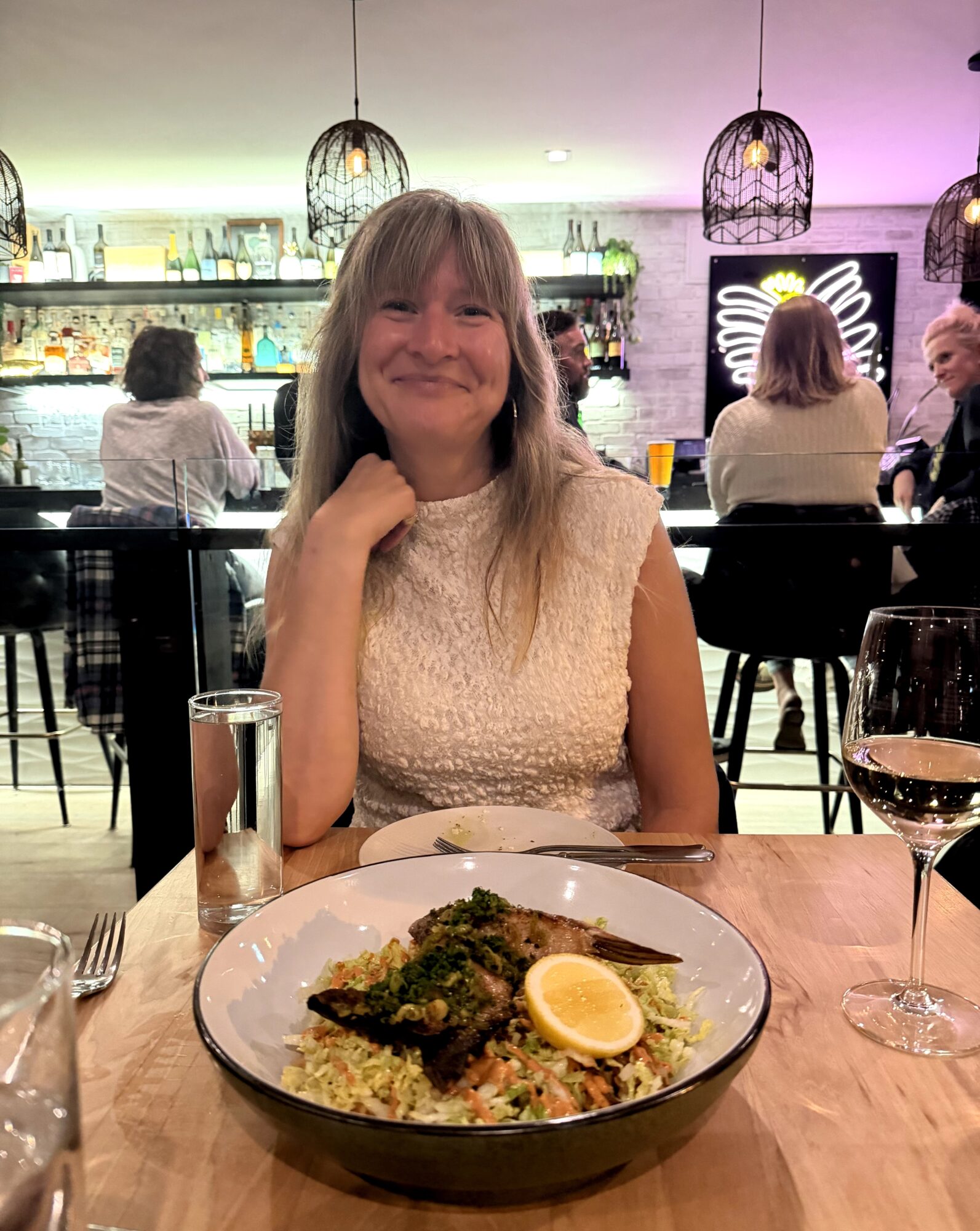 Woman smiling at a restaurant table with a plate of food, glass of wine, and a glass of water, in a modern setting.
