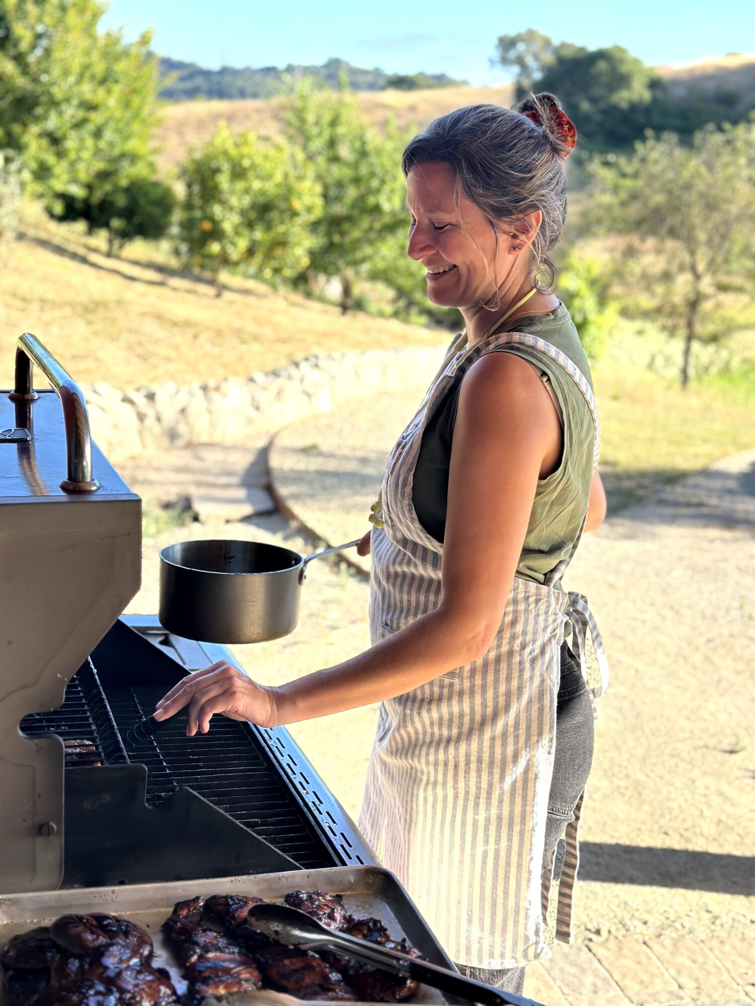 Woman grilling outdoors, smiling, wearing an apron, with trees and hills in the background.