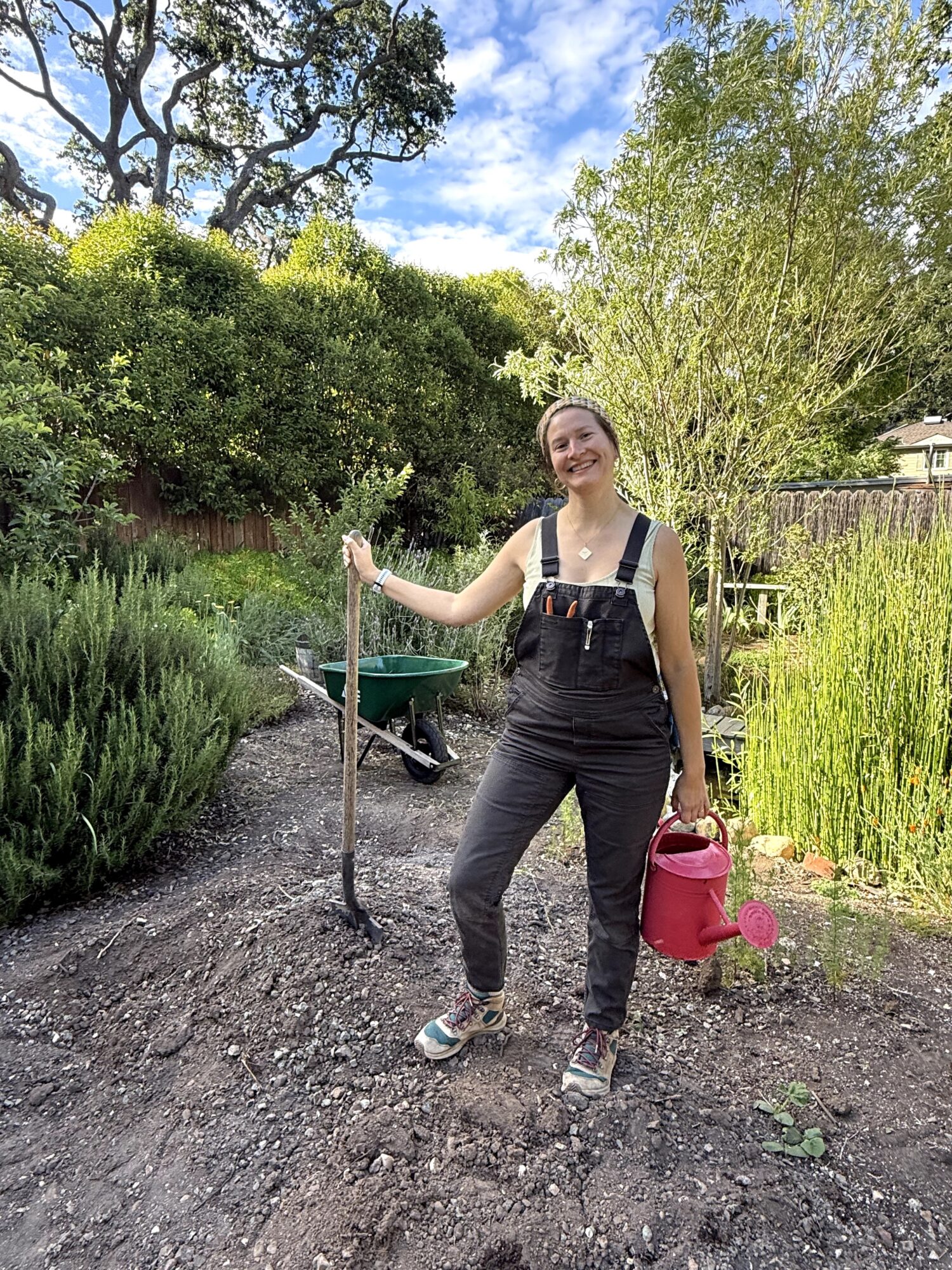 Young woman in black overalls watering plants in a garden with trees and bushes, smiling, holding a red watering can.