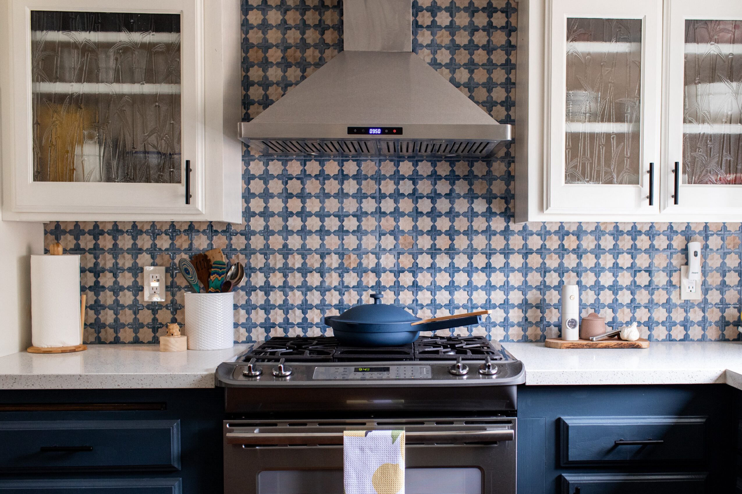 Kitchen with blue and white patterned backsplash, white upper cabinets, and dark lower cabinets, stove, and range hood.
