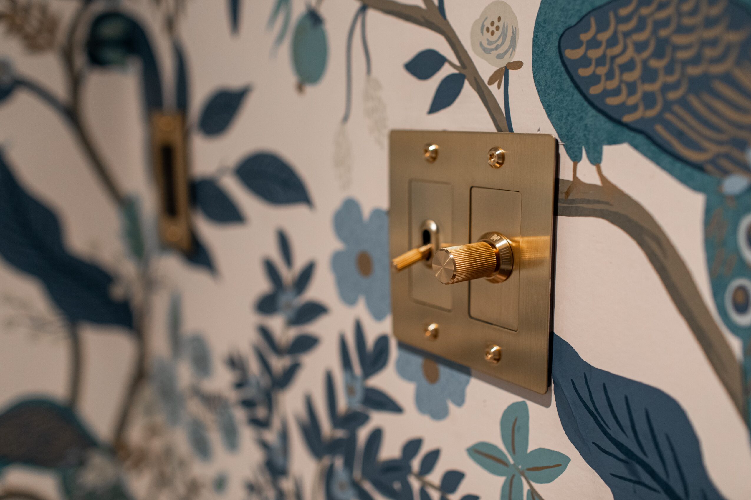 Switch on a wall with floral and bird wallpaper, gold-colored switch plate, and toggle switch. Word count: 20.