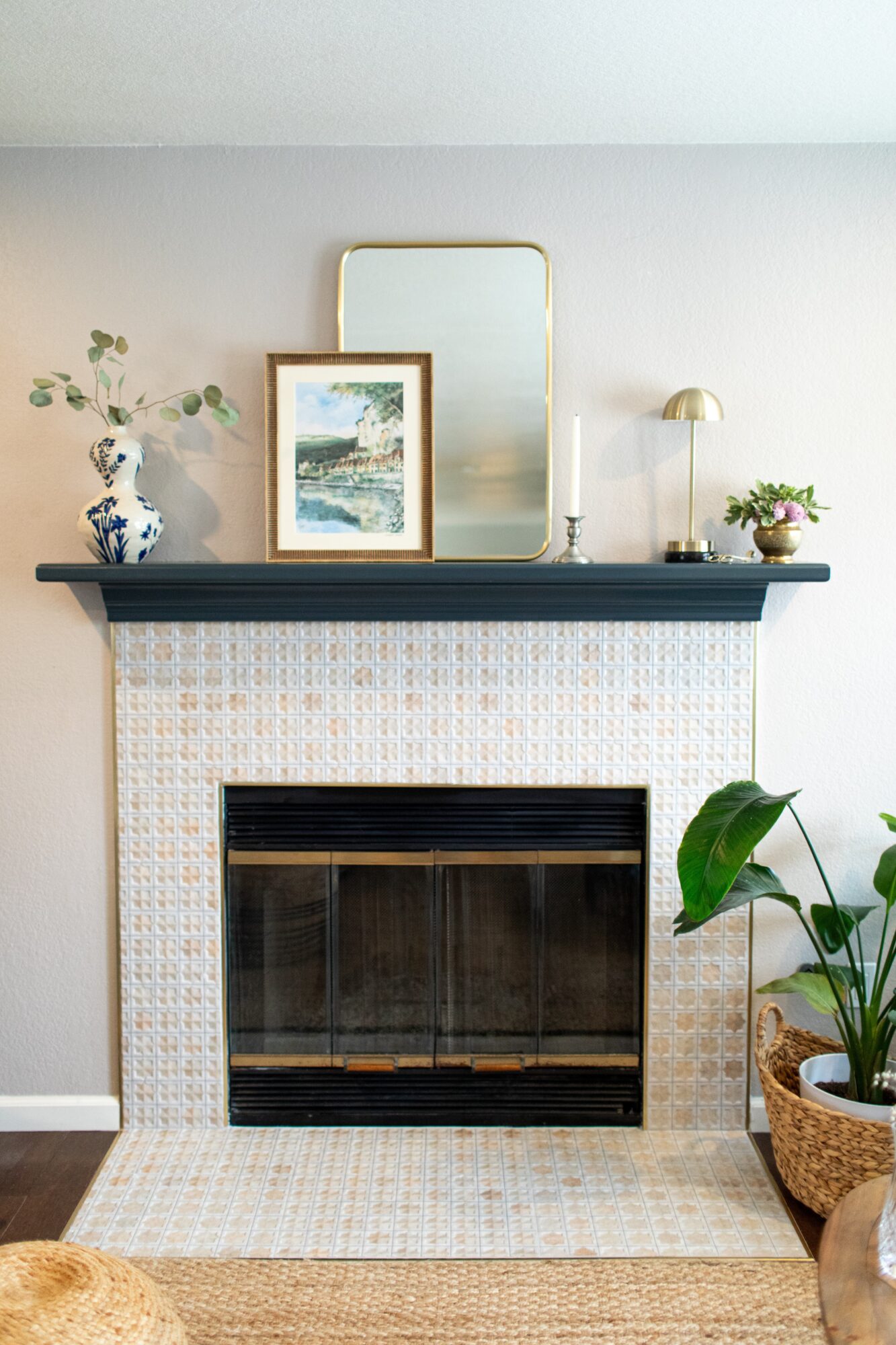 Fireplace with a dark mantel, decorated with a framed picture, mirror, candle, and plants, against a light wall.