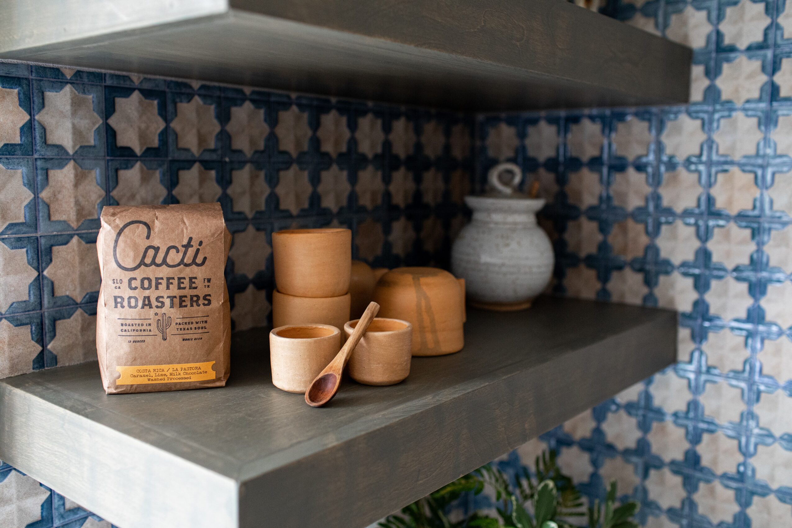 Shelf with coffee cups, a spoon, a bag of coffee, and a ceramic container against a patterned blue and white tiled wall.