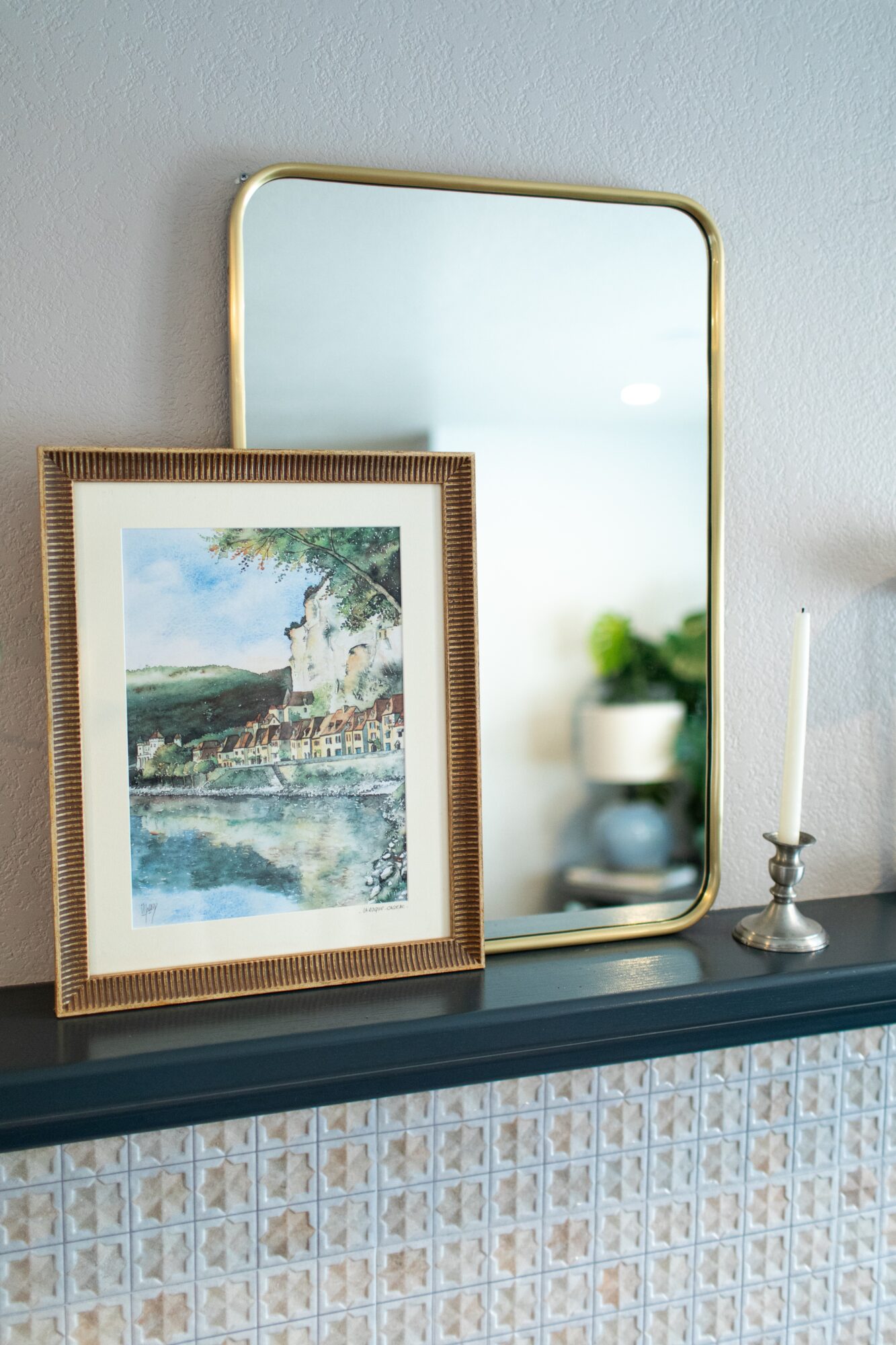 Decorative mirror and framed picture on a dark shelf with a candle holder and plant, against a light wall.