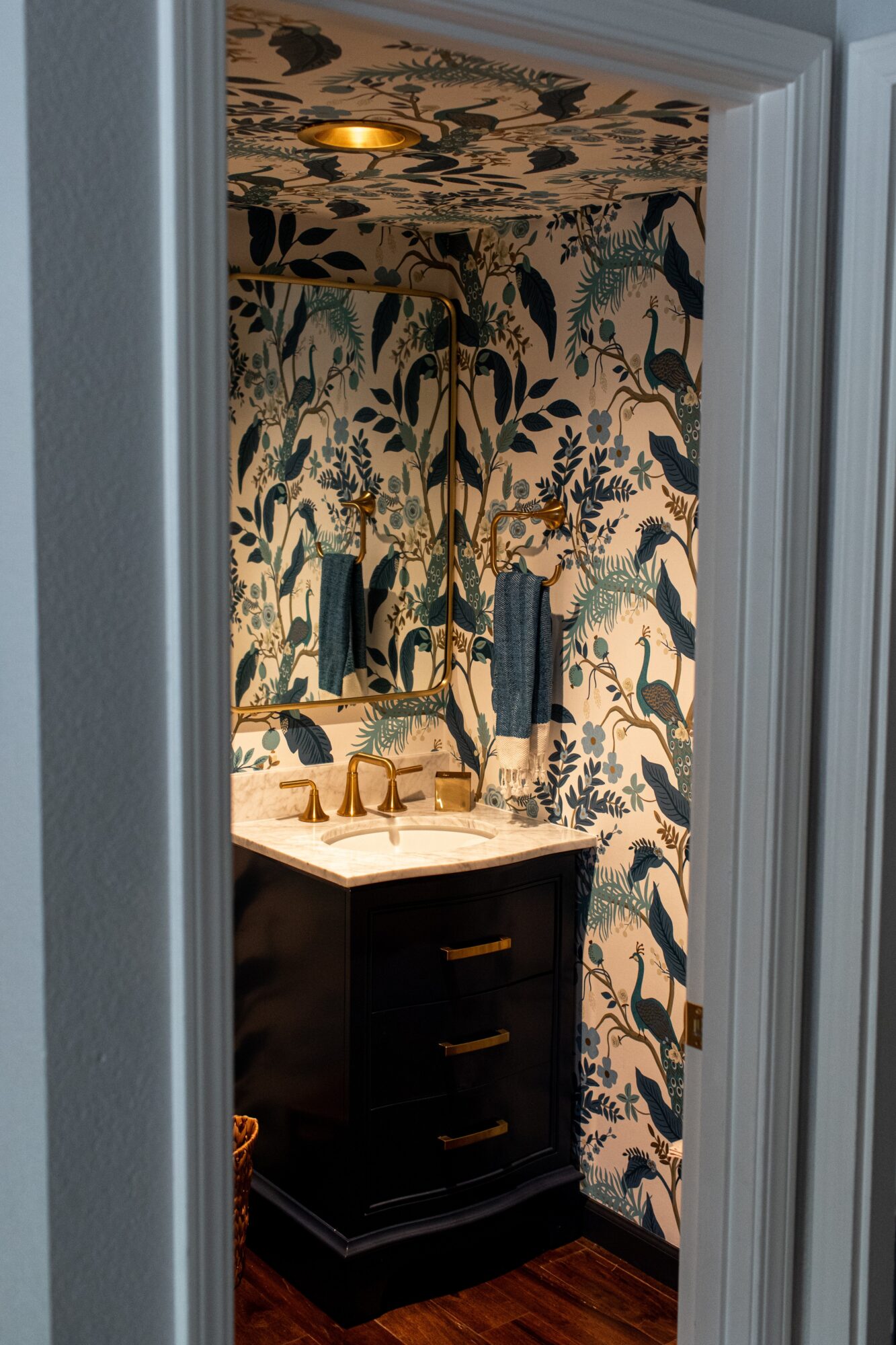 Bathroom with patterned wallpaper, dark vanity with gold handles, and a mirror, viewed through a doorway.