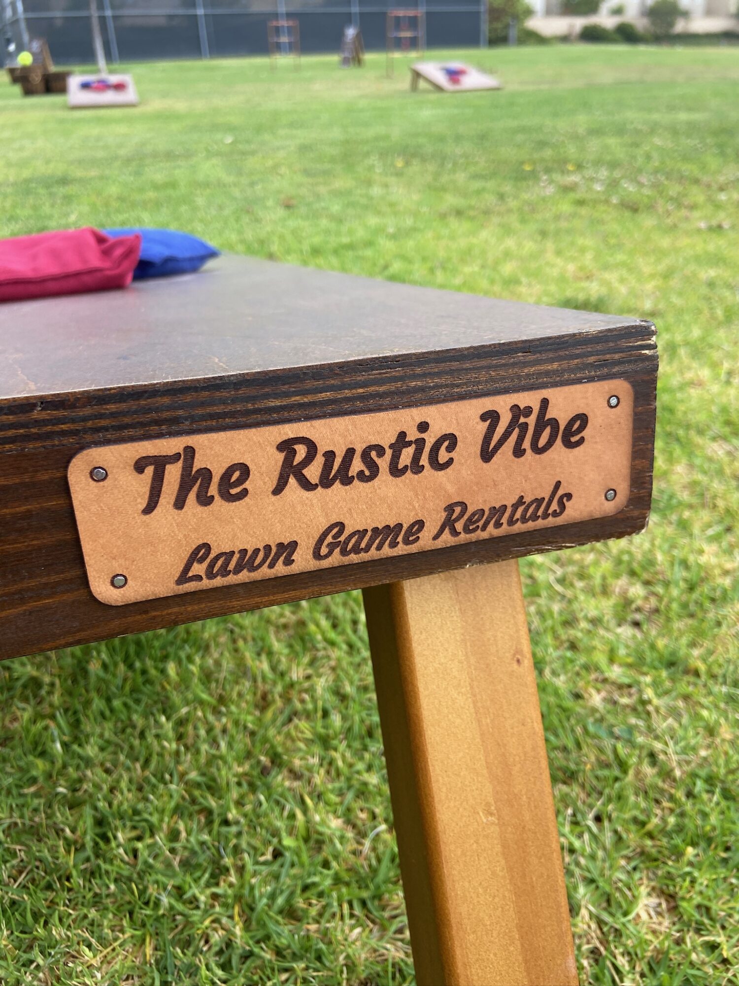 Close-up of a wooden sign on a table reading 'The Rustic Vibe Lawn Game Rentals' in a grassy outdoor area.