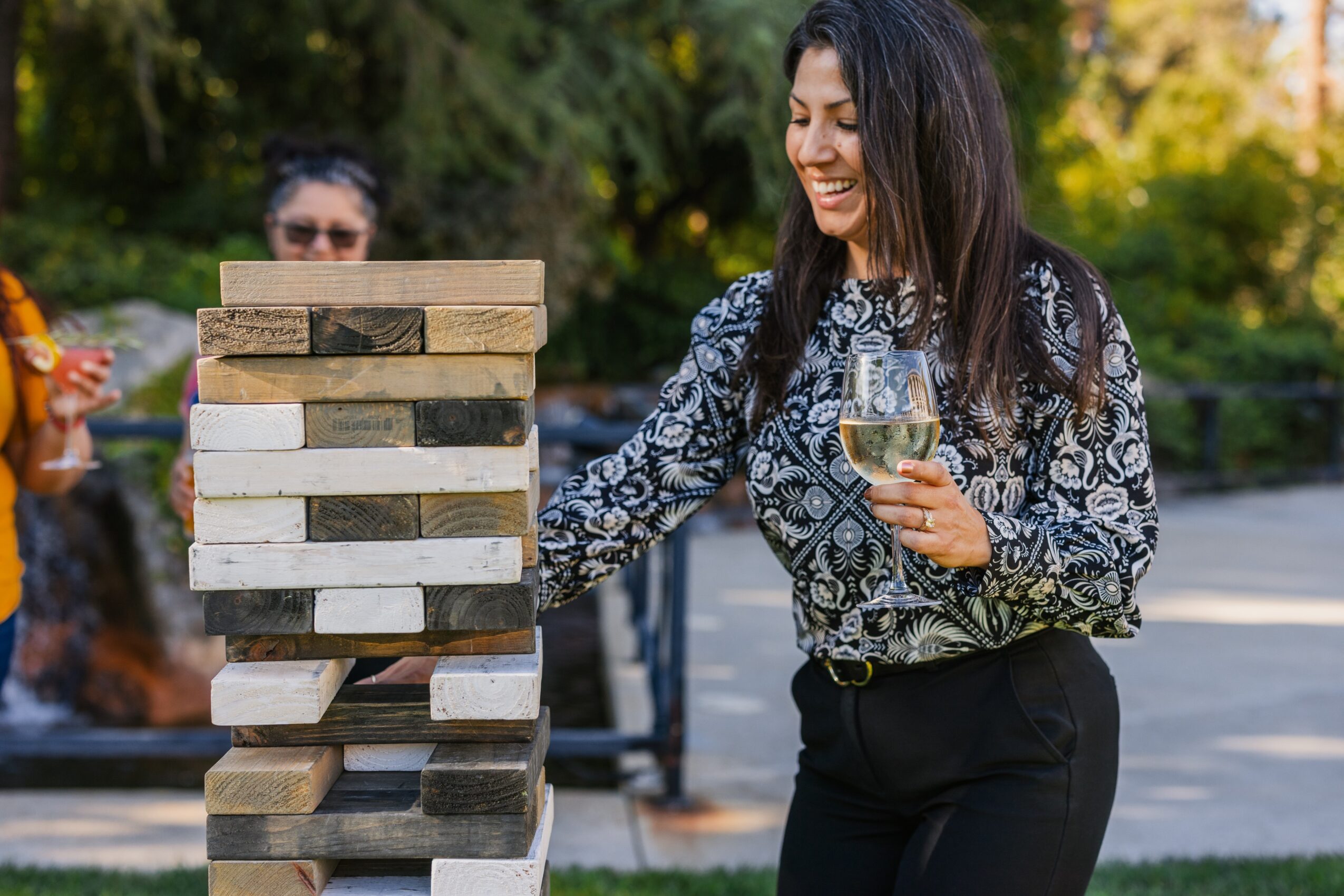 Woman playing giant Jenga outdoors, holding a glass of wine, smiling, with trees and a person in the background.