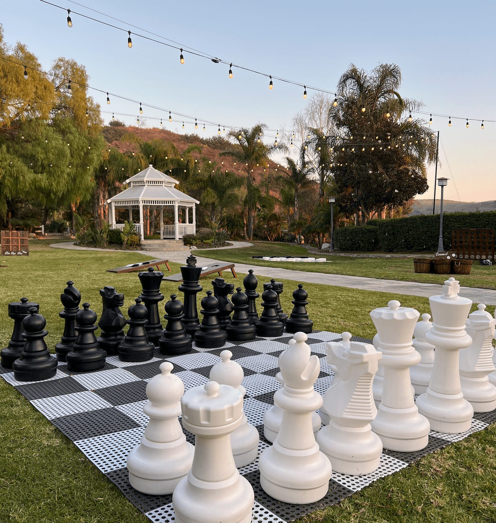 Large outdoor chess set with black and white pieces on a checkered mat in a park with trees and string lights.