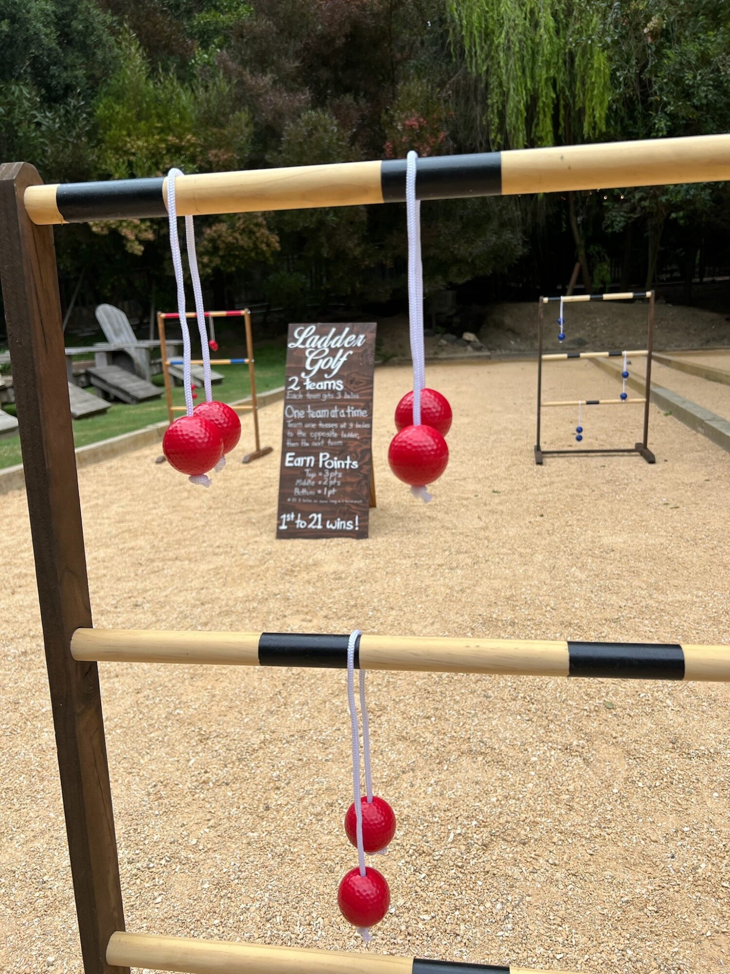 Three red balls hanging from a horizontal bar with a sign in the background, outdoor setting with trees and a sandy ground.