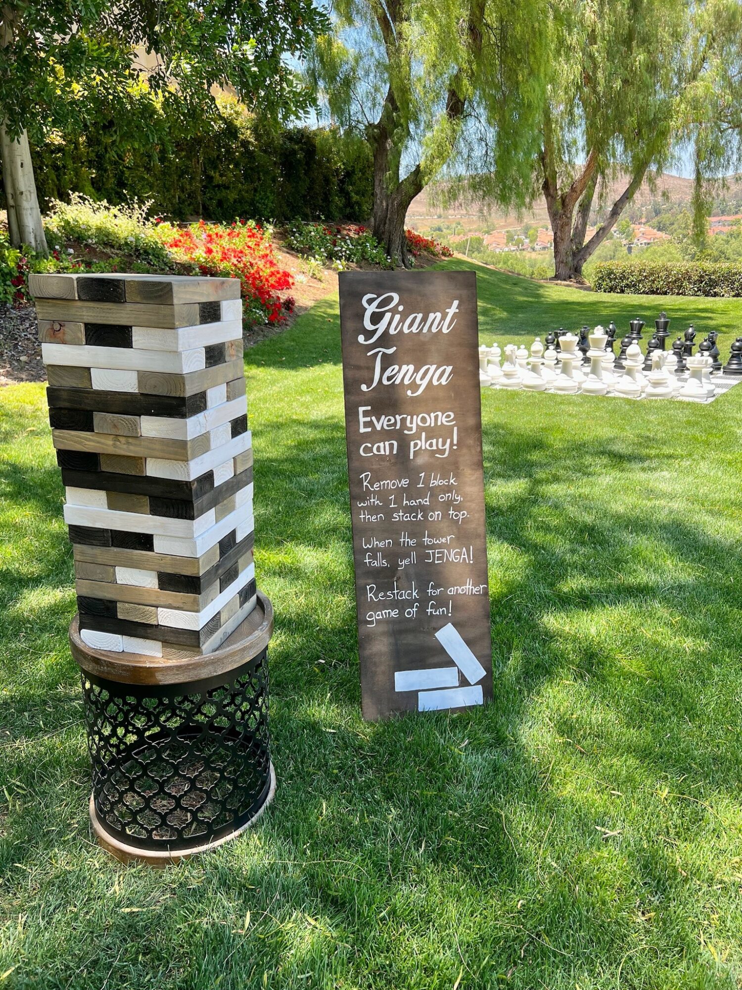 Signboard for giant Jenga game outdoors, with a wooden Jenga tower and chess pieces on grass, trees, and flowers in background.