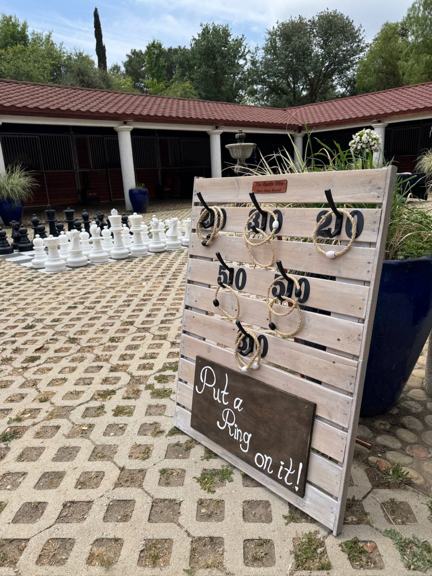 Outdoor courtyard with large chess set, decorative board with hooks, and a sign reading 'Put a ring on it!'.