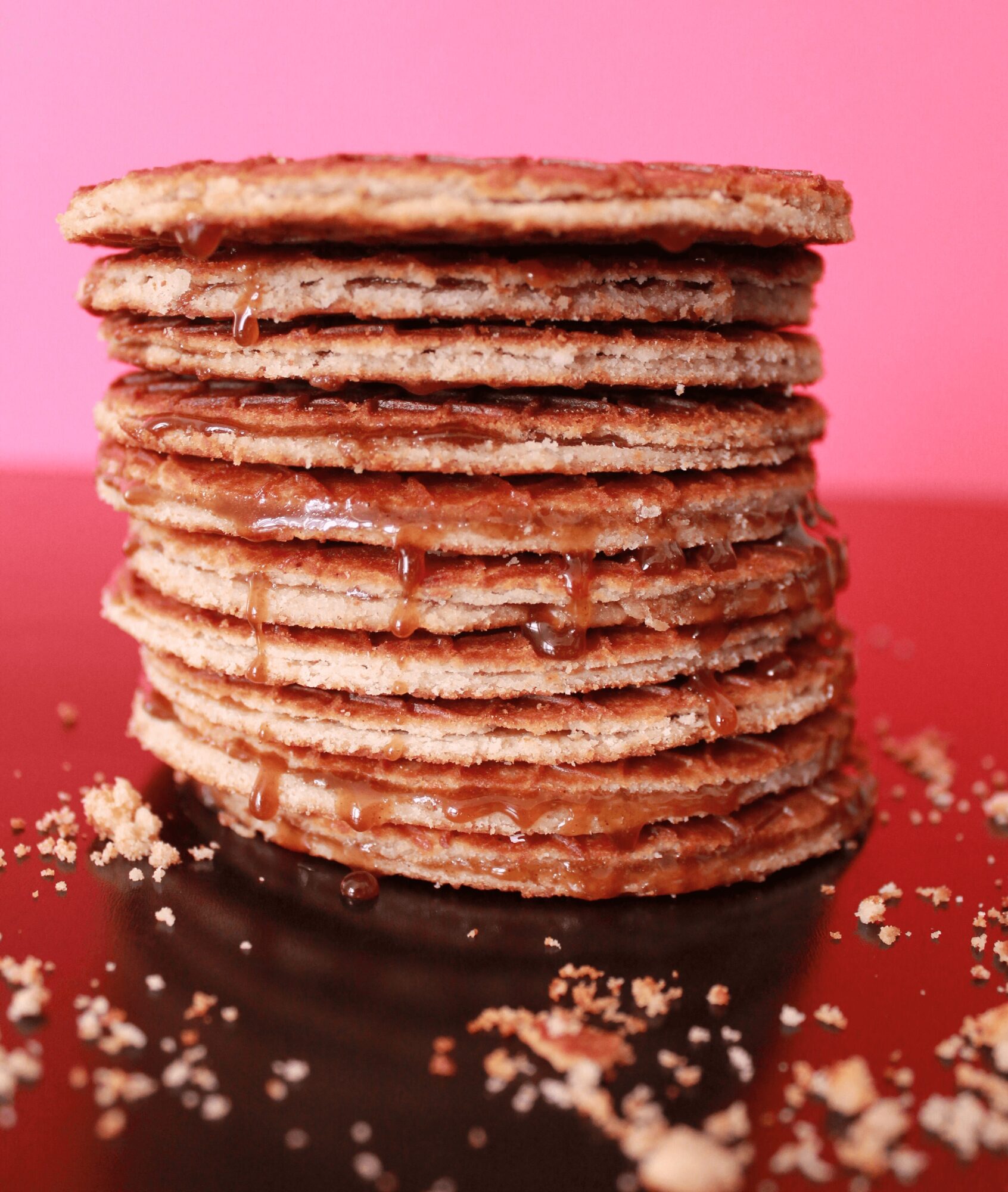Stack of multiple thin, round cookies with a glossy filling, on a black surface with crumbs, pink background.