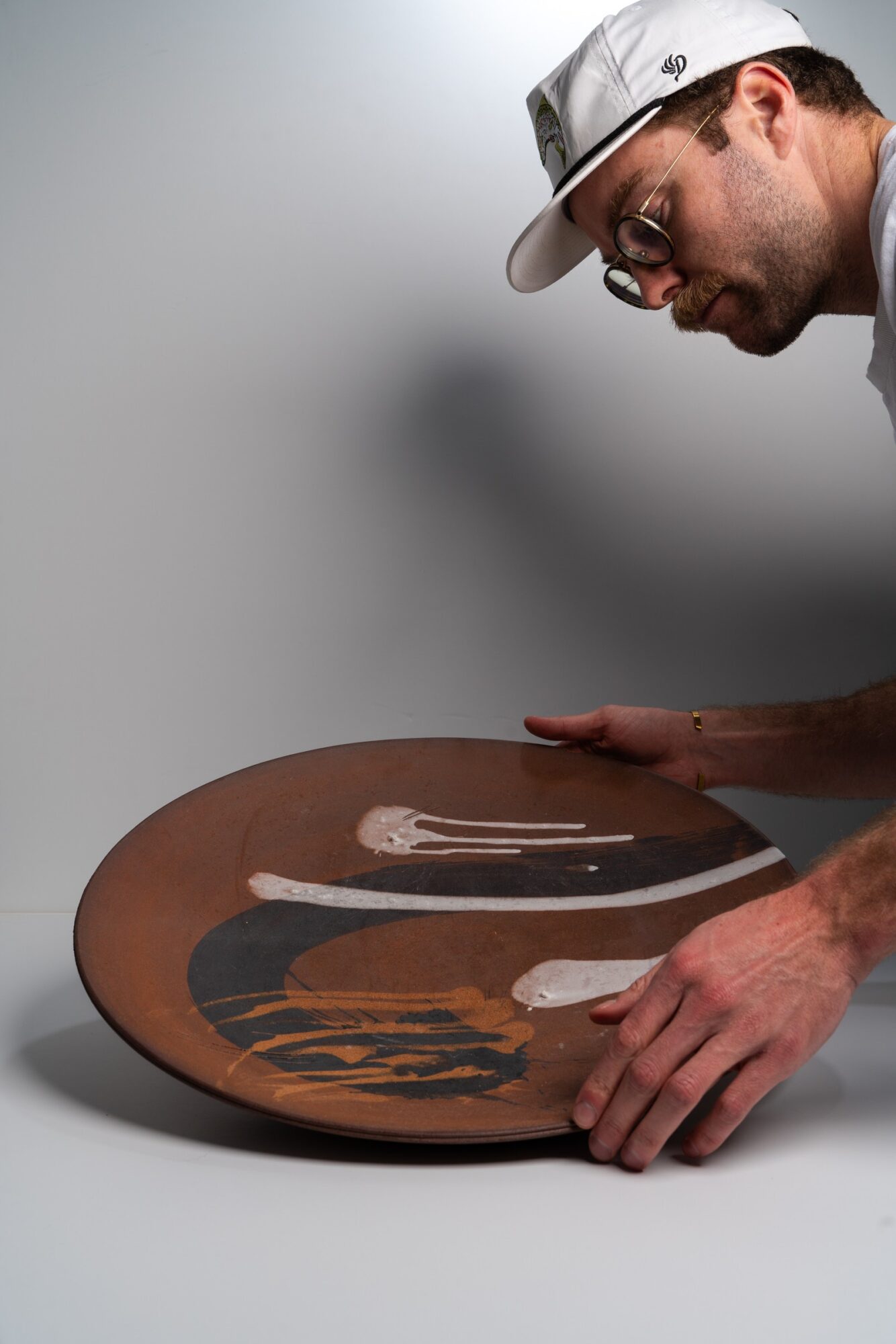 Person wearing glasses and a cap looks at a large, decorated ceramic plate on a table.