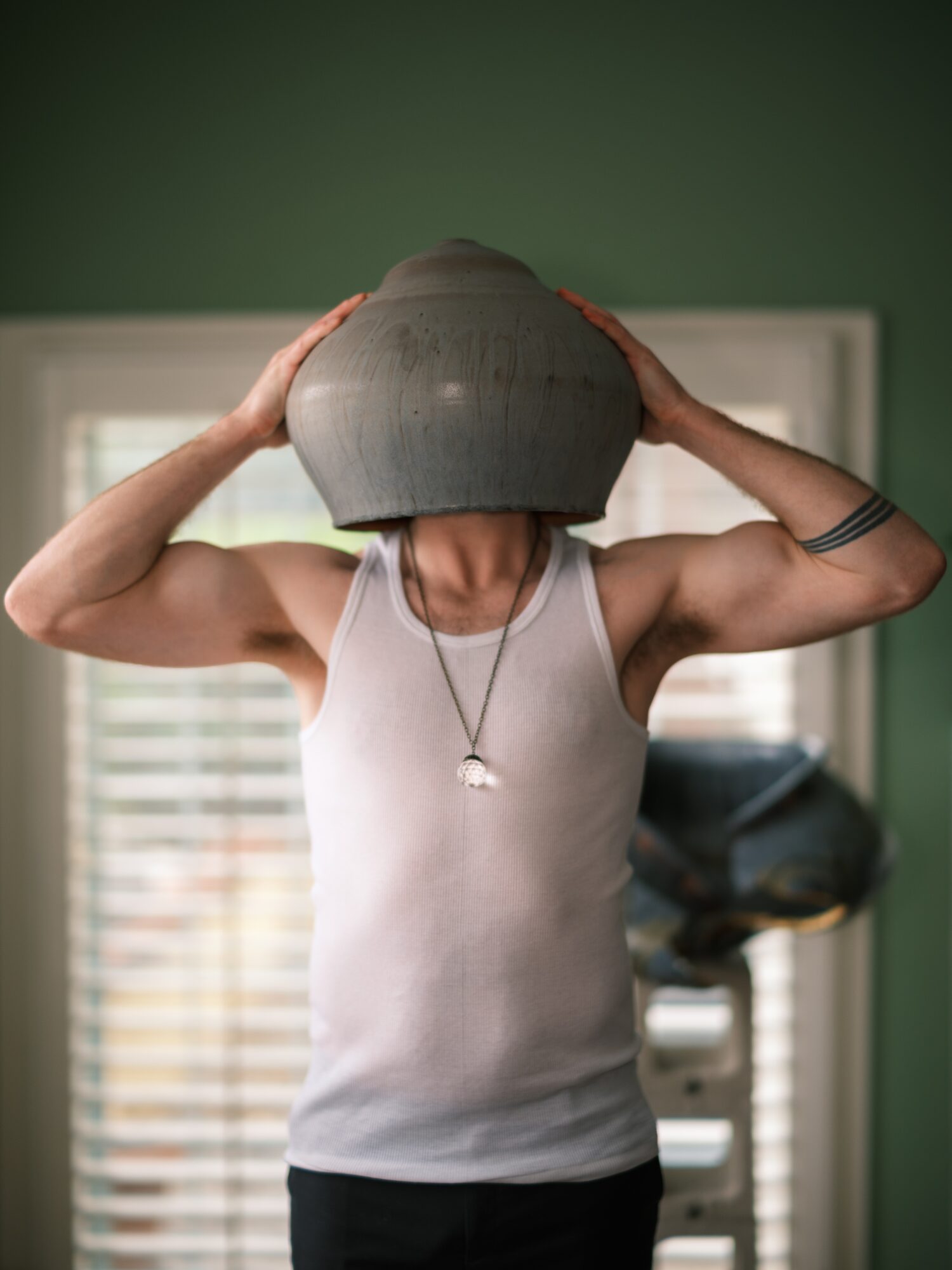 Person holding a large gray object over their head, wearing a white tank top and a necklace, in a room with green walls.