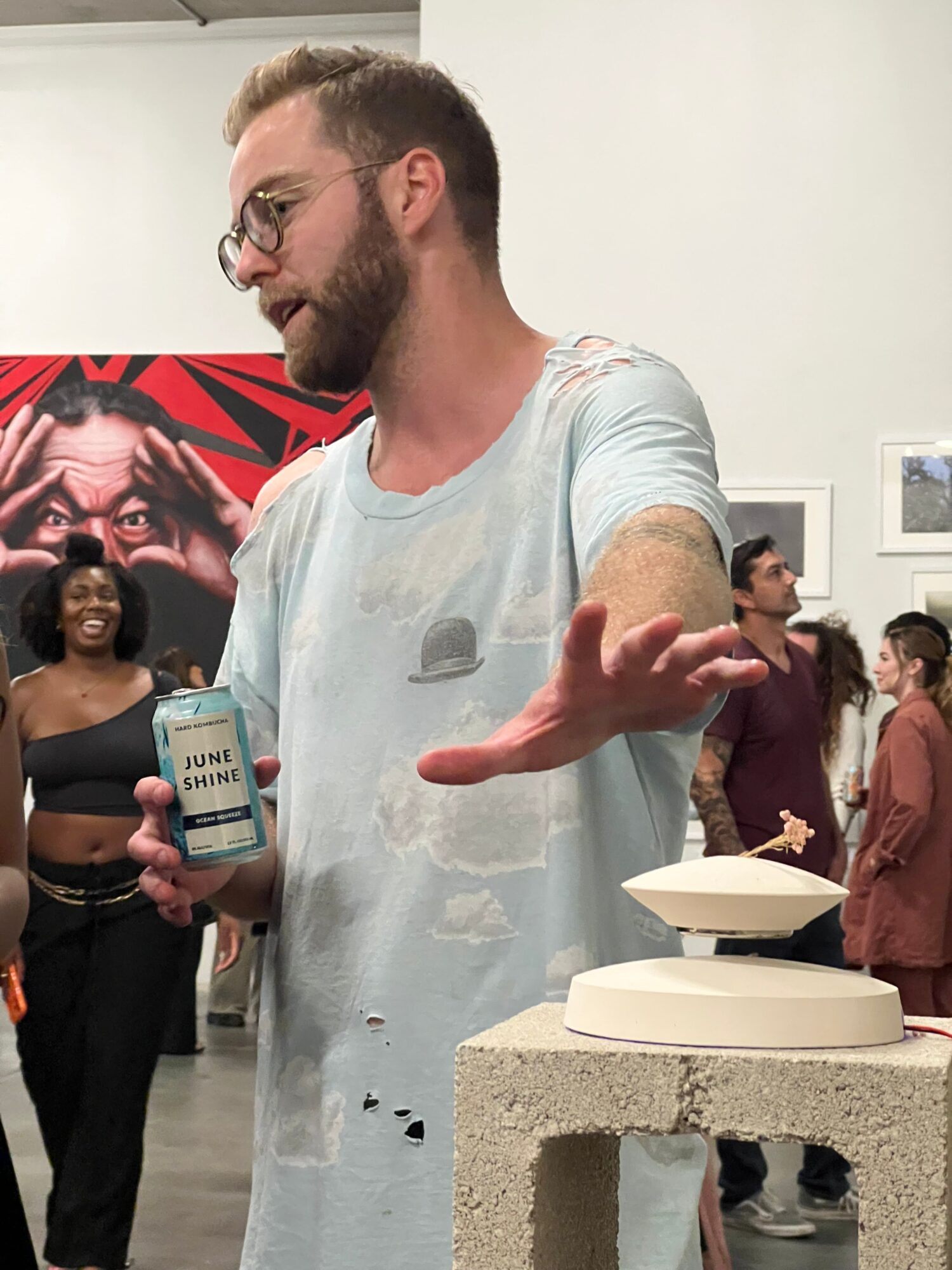 Man with glasses and beard wearing a plastic apron, holding a phone, in a room with people and artwork in the background.